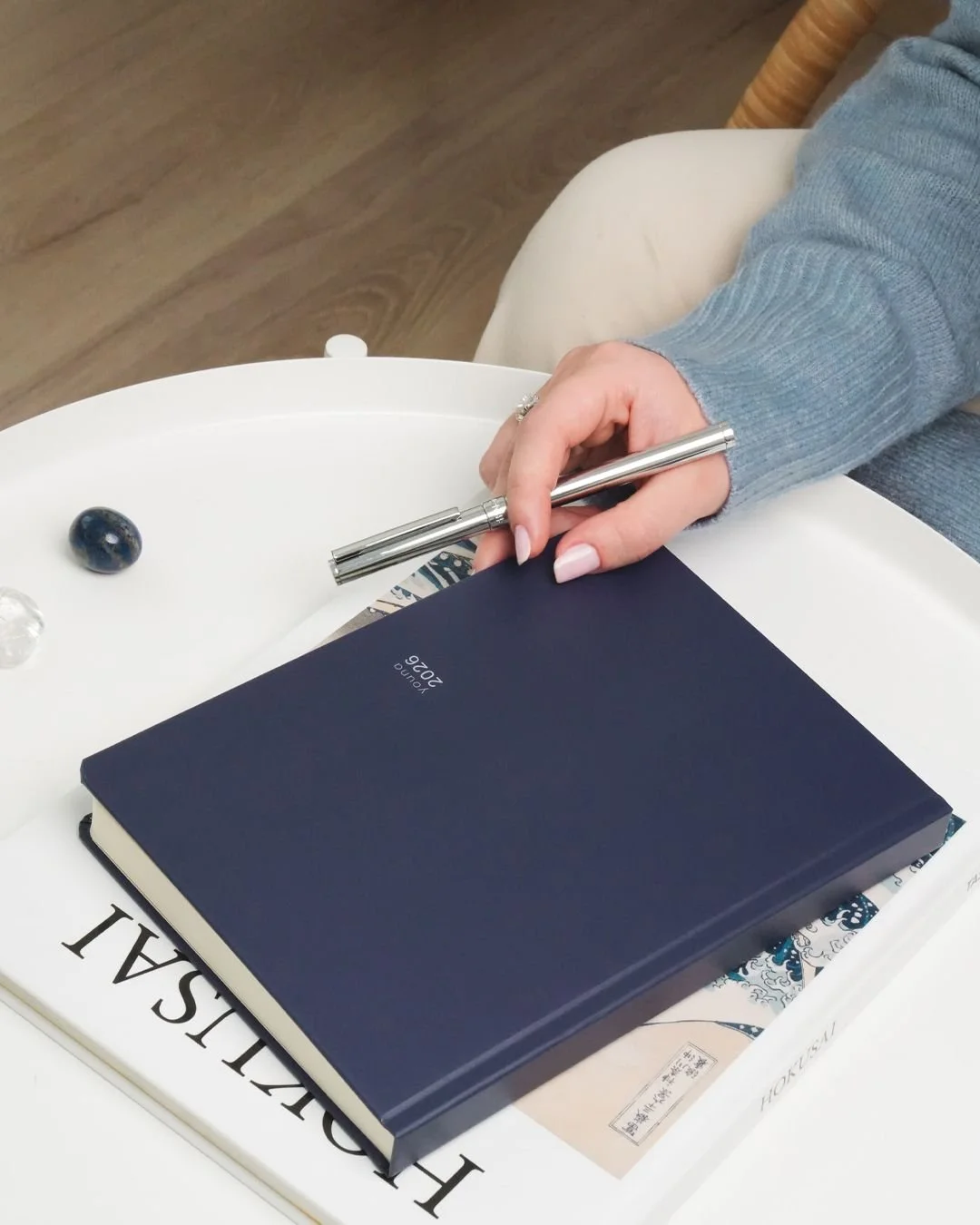 A person with a light blue sweater holding a silver pen, sitting near a white tray with a dark blue planner, a navy marble, a clear glass marble, and a book titled 'HISTORICAL'.