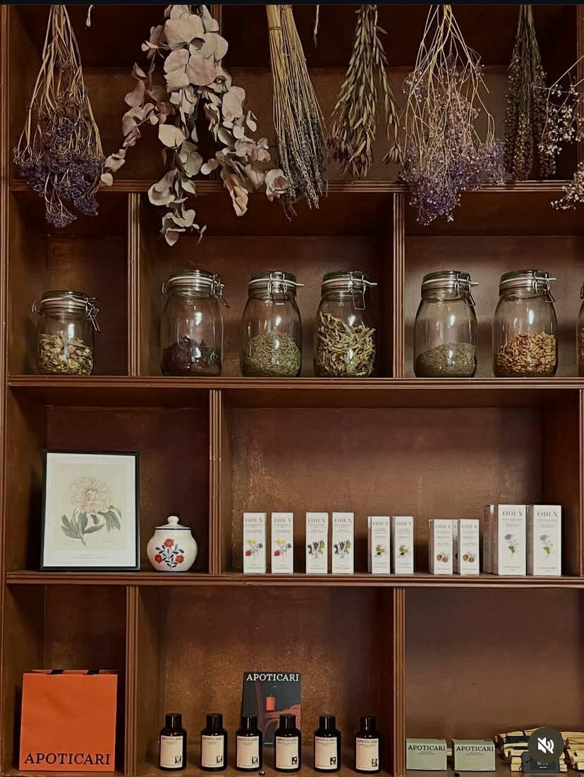 Étagère en bois avec des bocaux en verre remplis de plantes séchées, des boîtes de produits apoticar, un cadre avec une illustration de fleur, et des petits flacons de parfum ou huile artisanale, accompagnés de bouquets d'herbes suspendus en haut.