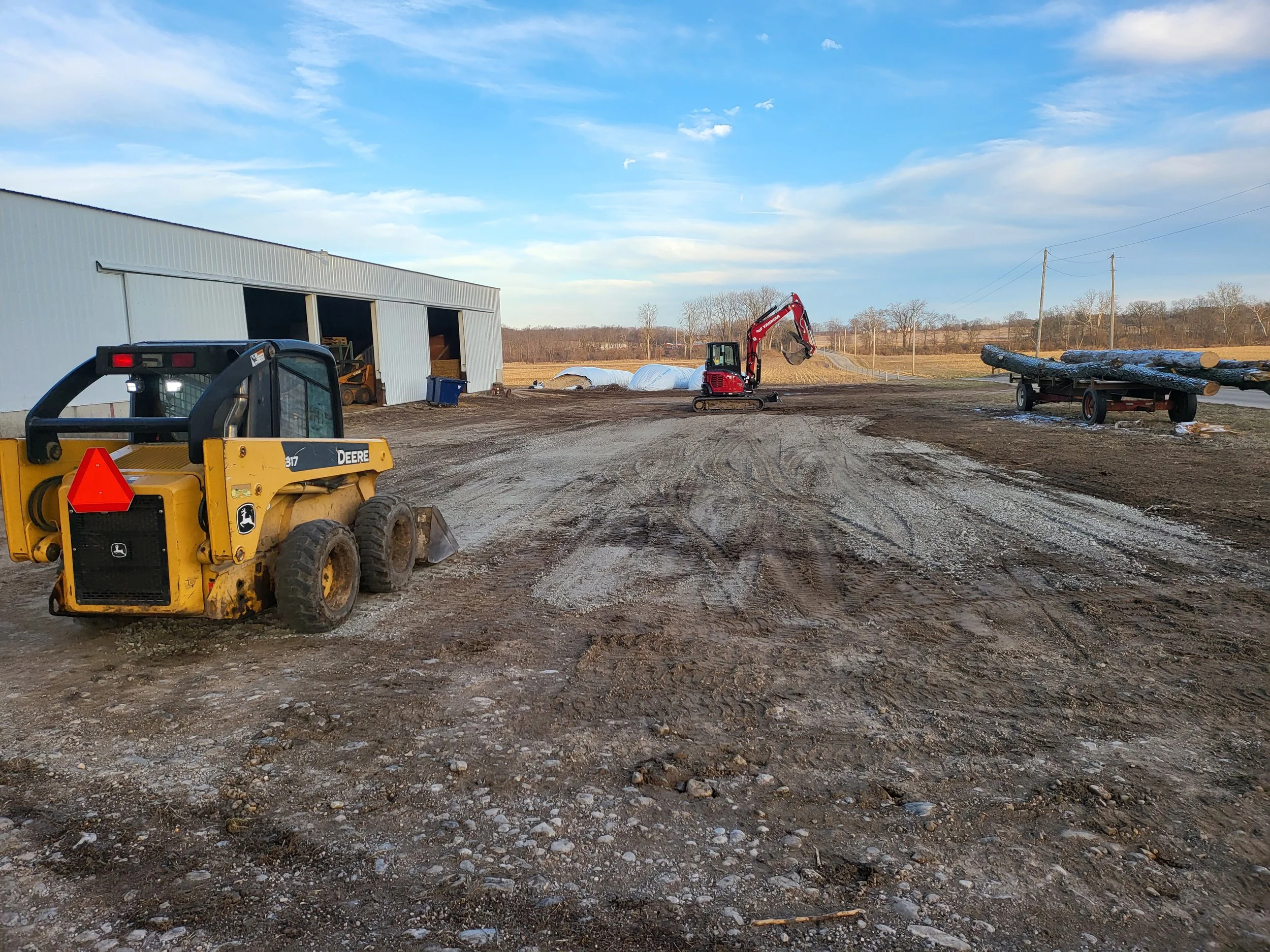 A construction site with a yellow John Deere mini excavator, a red mini excavator, and logs on a trailer in front of a large metal building on a gravel lot under a partly cloudy sky.