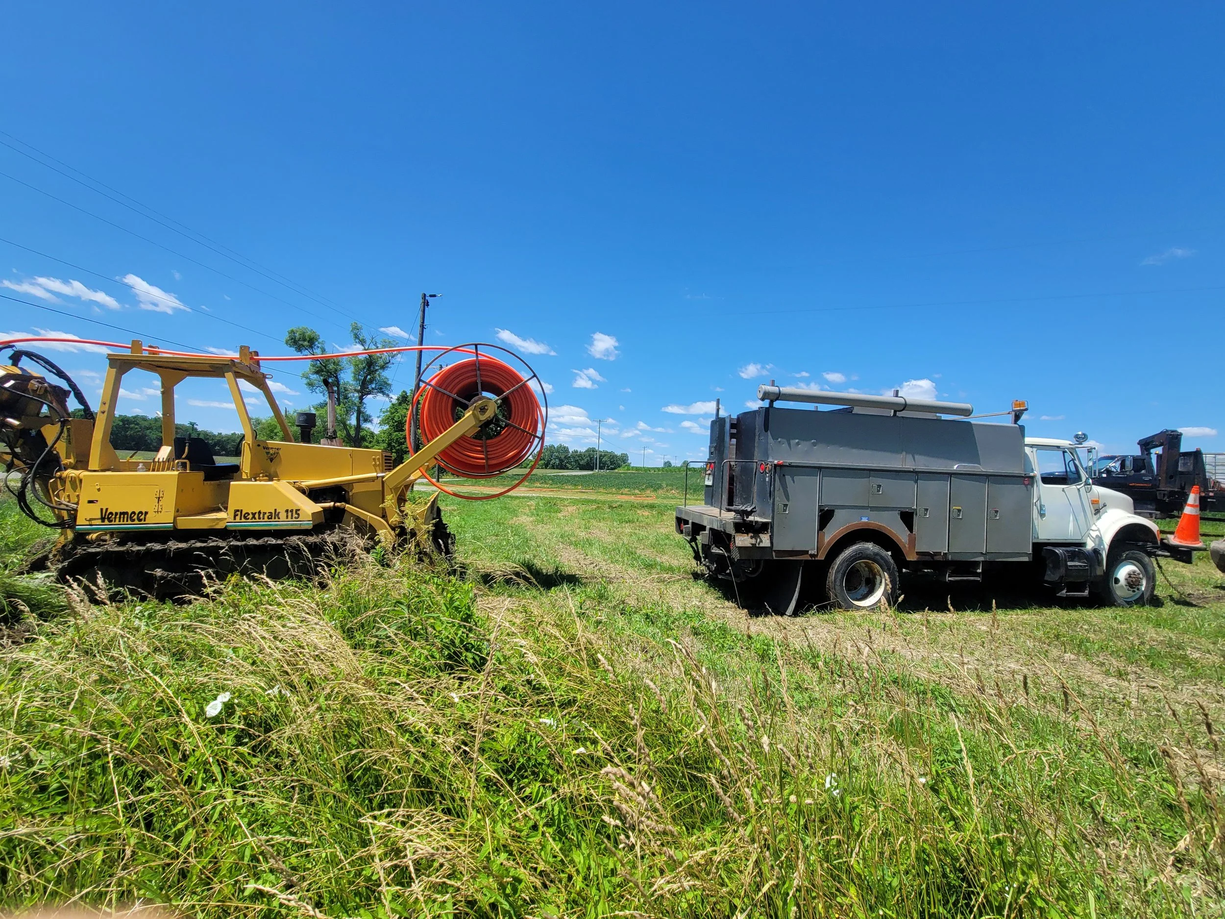 Farm field with yellow Vermeer Flextrax 115 turf tiller and a gray utility truck on bright sunny day with blue sky and green grass.