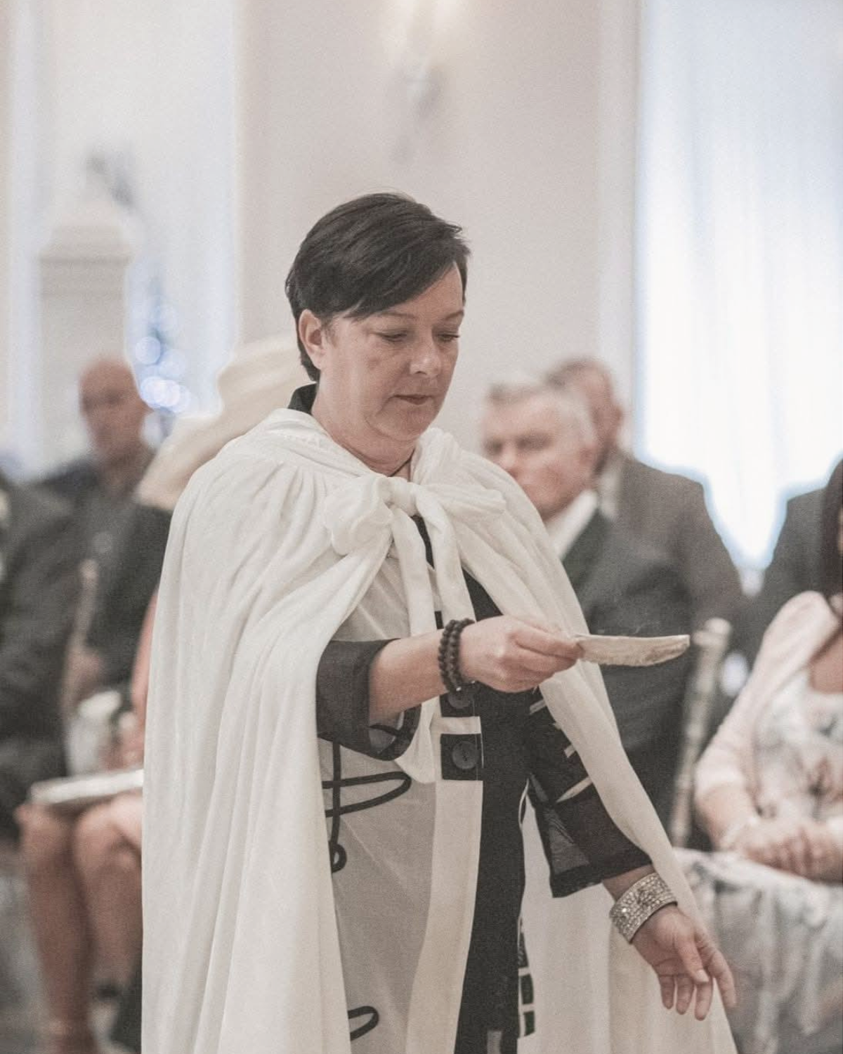 A woman in a black and white outfit, draped with a white cape, standing and reading from a paper in a room with other seated people.