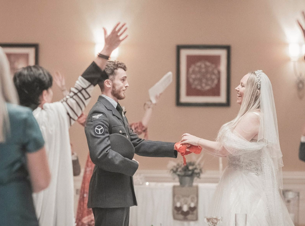 A bride and groom holding hands during their wedding ceremony, with guests raising their hands in blessing or celebration.