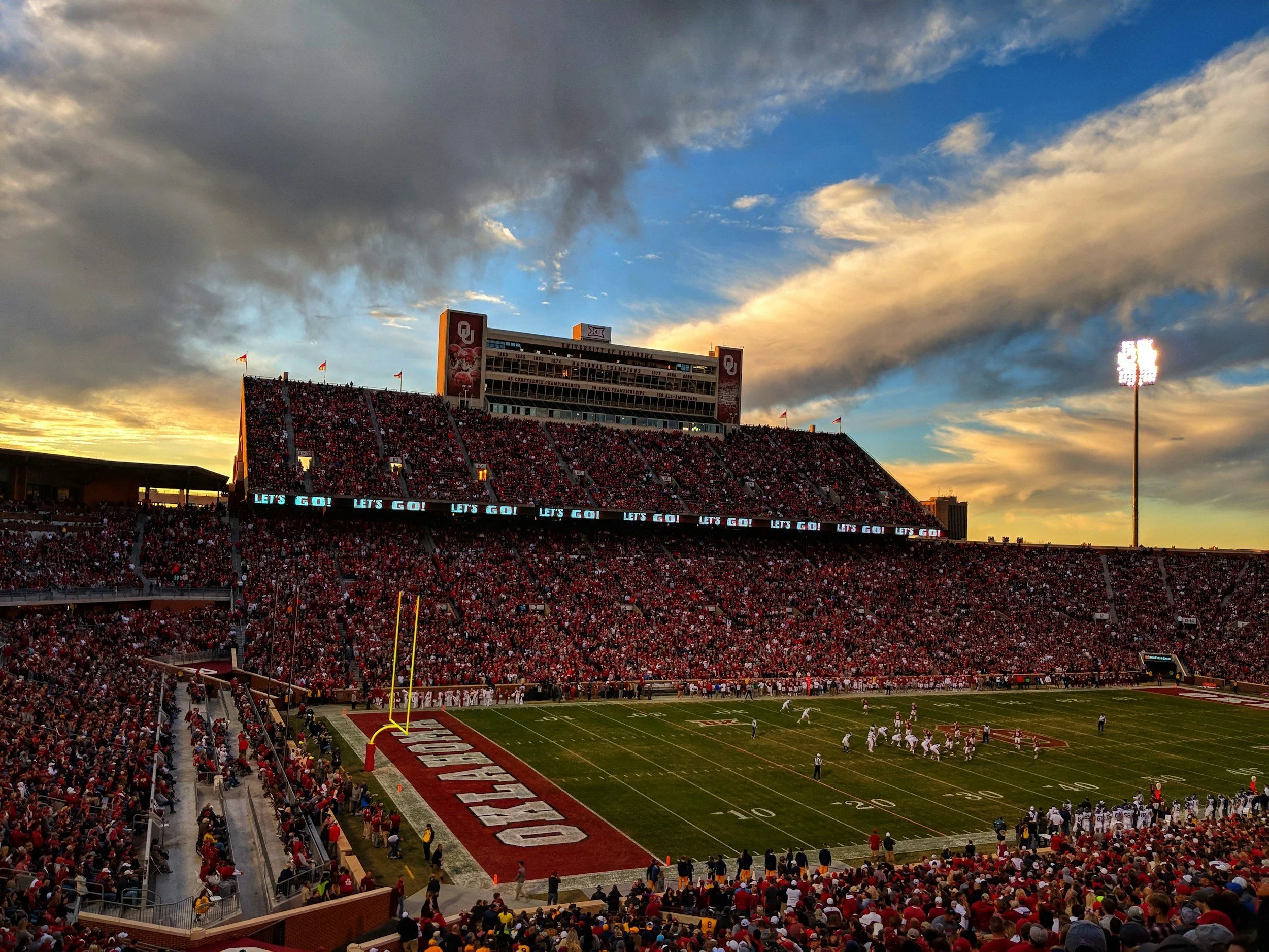 OU football stadium during sunset with fans in the stands, players on the field, and large stadium lights illuminating the field.