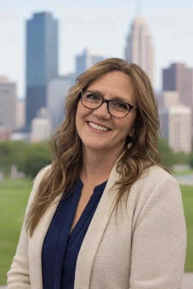 Smiling woman with glasses and wavy hair outdoors with city skyline in the background.