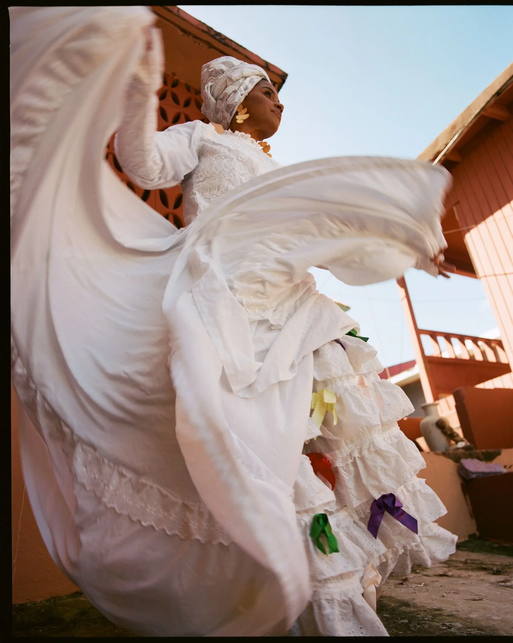 Mujer vestida con ropa blanca y sombrero, bailando con faldas de encaje y lazos de colores en un entorno exterior