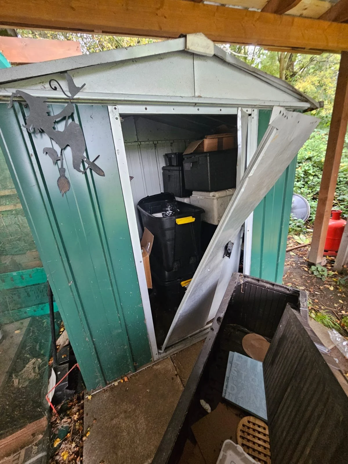 A small green shed with a white door partially open, revealing storage boxes, plastic containers, and tools inside. A metal witch silhouette decoration is mounted outside near the shed.