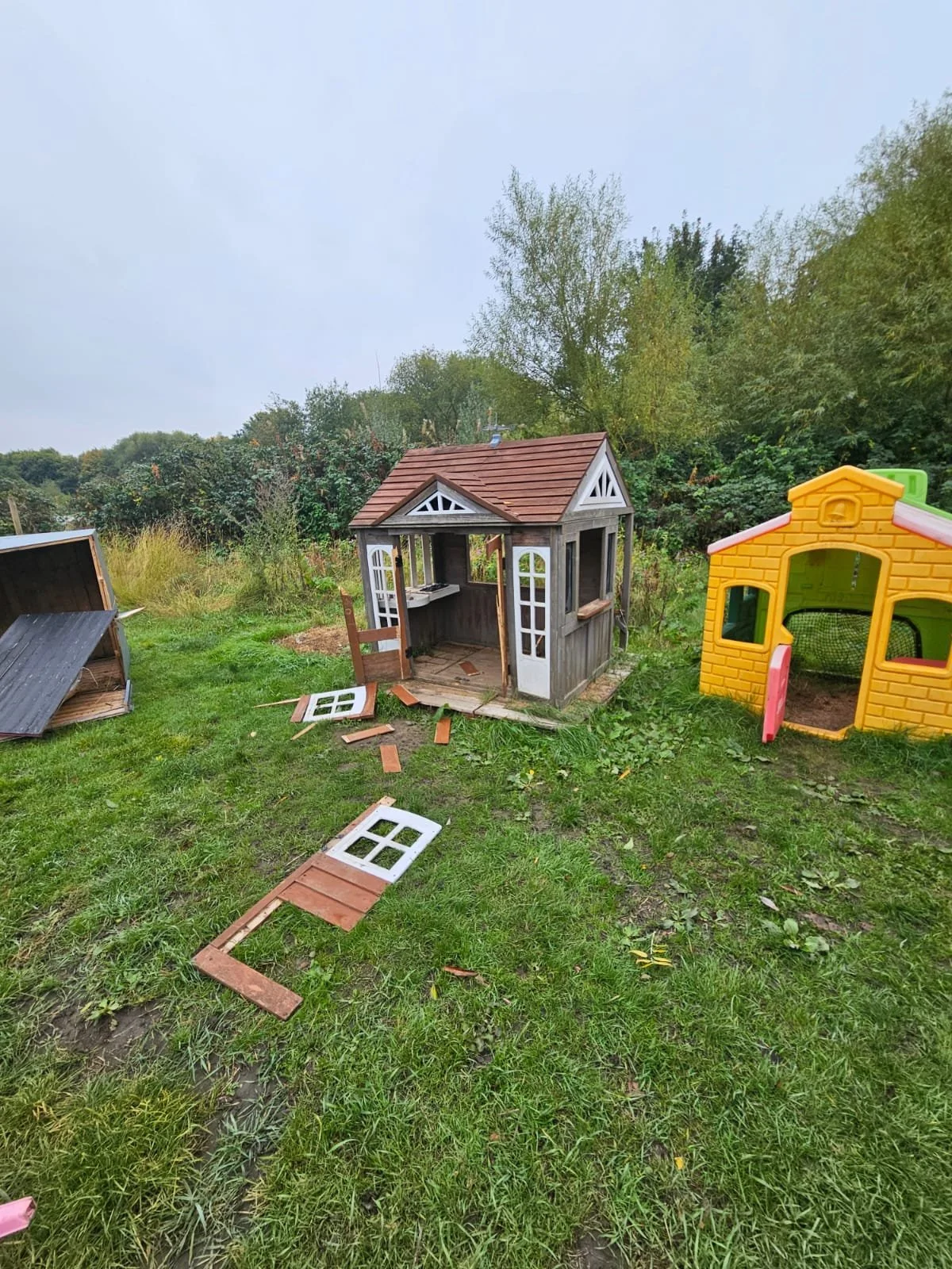 Children’s toy playhouses, one black and white with a red roof, another yellow with a pink door, on a grassy yard, some parts broken and scattered.