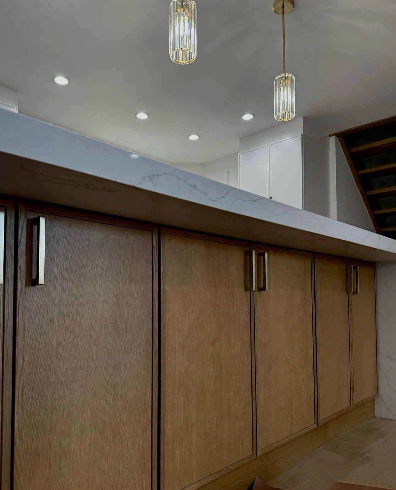 View of a kitchen counter with wooden cabinet doors and a marble countertop, with ceiling lights and hanging decorative pendant lights.