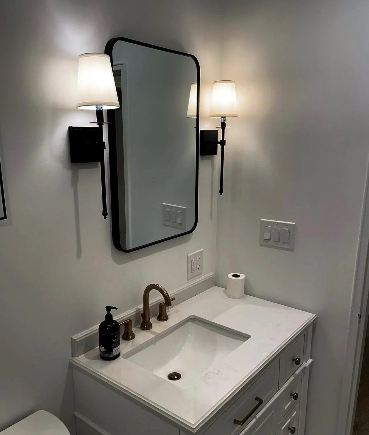 Bathroom vanity with white countertop, bronze faucet, mirror, wall-mounted light fixtures, soap dispenser, and a roll of toilet paper.