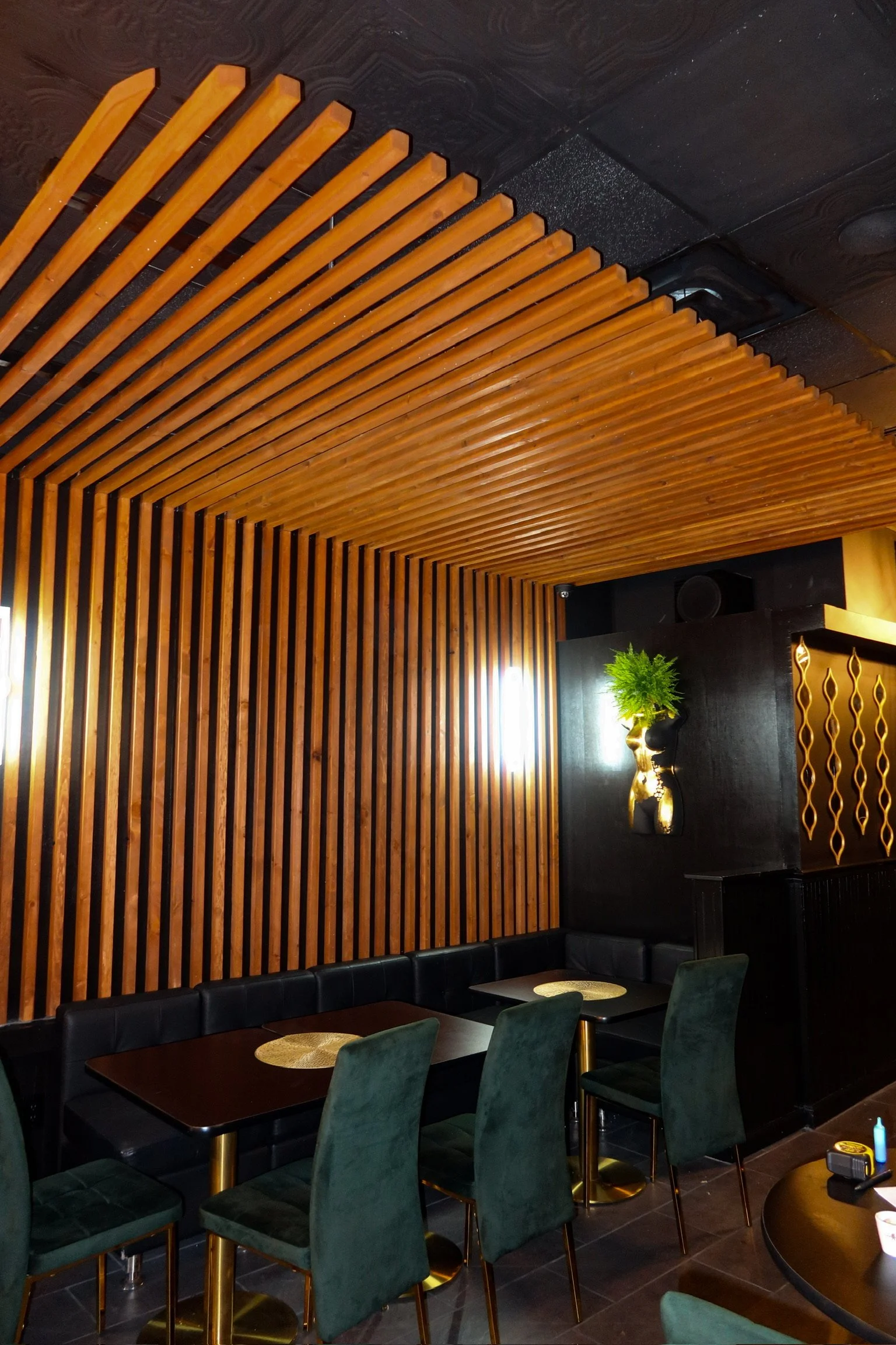 Interior of a restaurant or cafe with wooden slat wall and ceiling, black cushioned seating, dark tables with gold bases, and a decorative vase with green plant on a black partition wall.