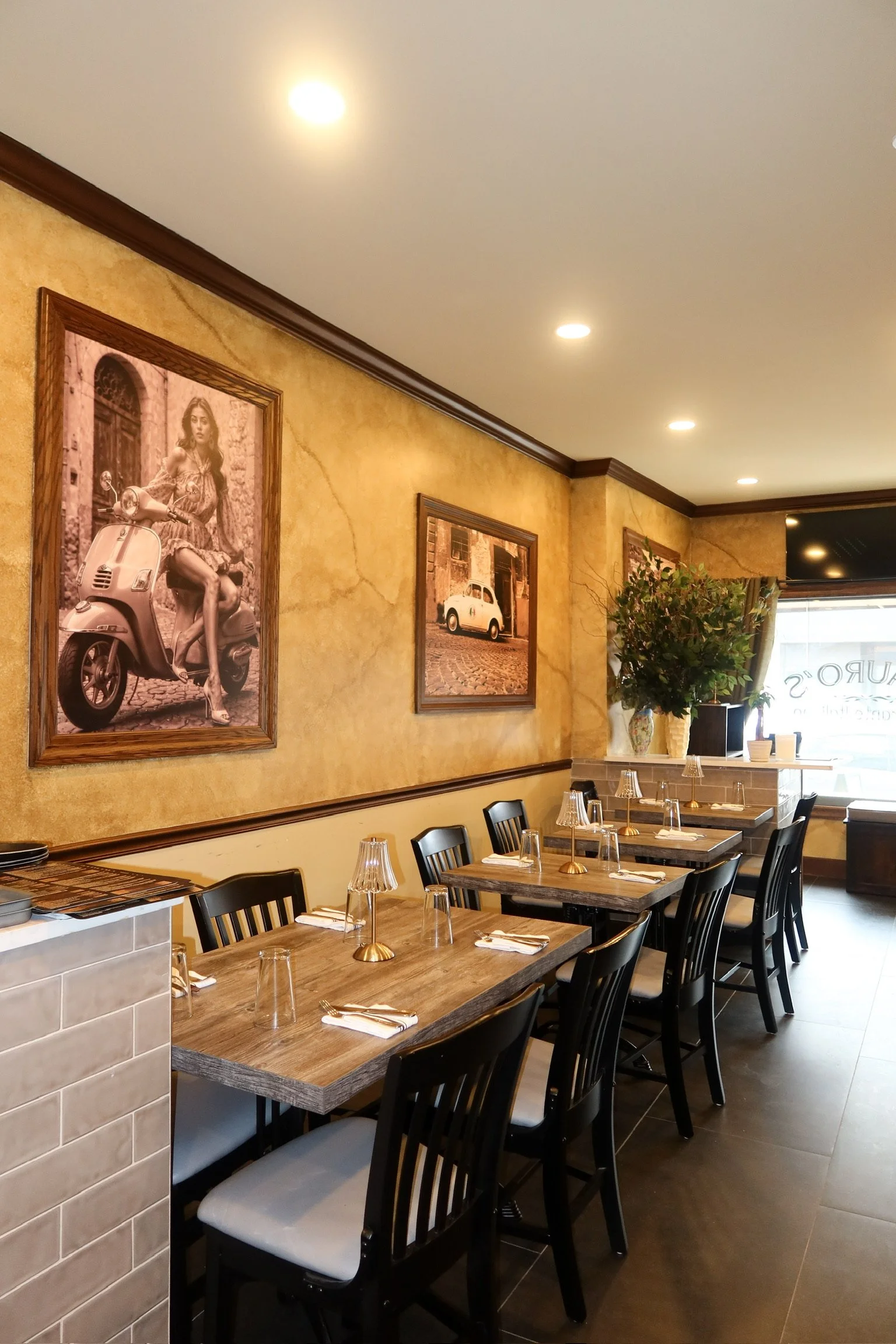 Interior of a restaurant with a row of wooden tables and black chairs, wall art with vintage photos, and a large window at the front.