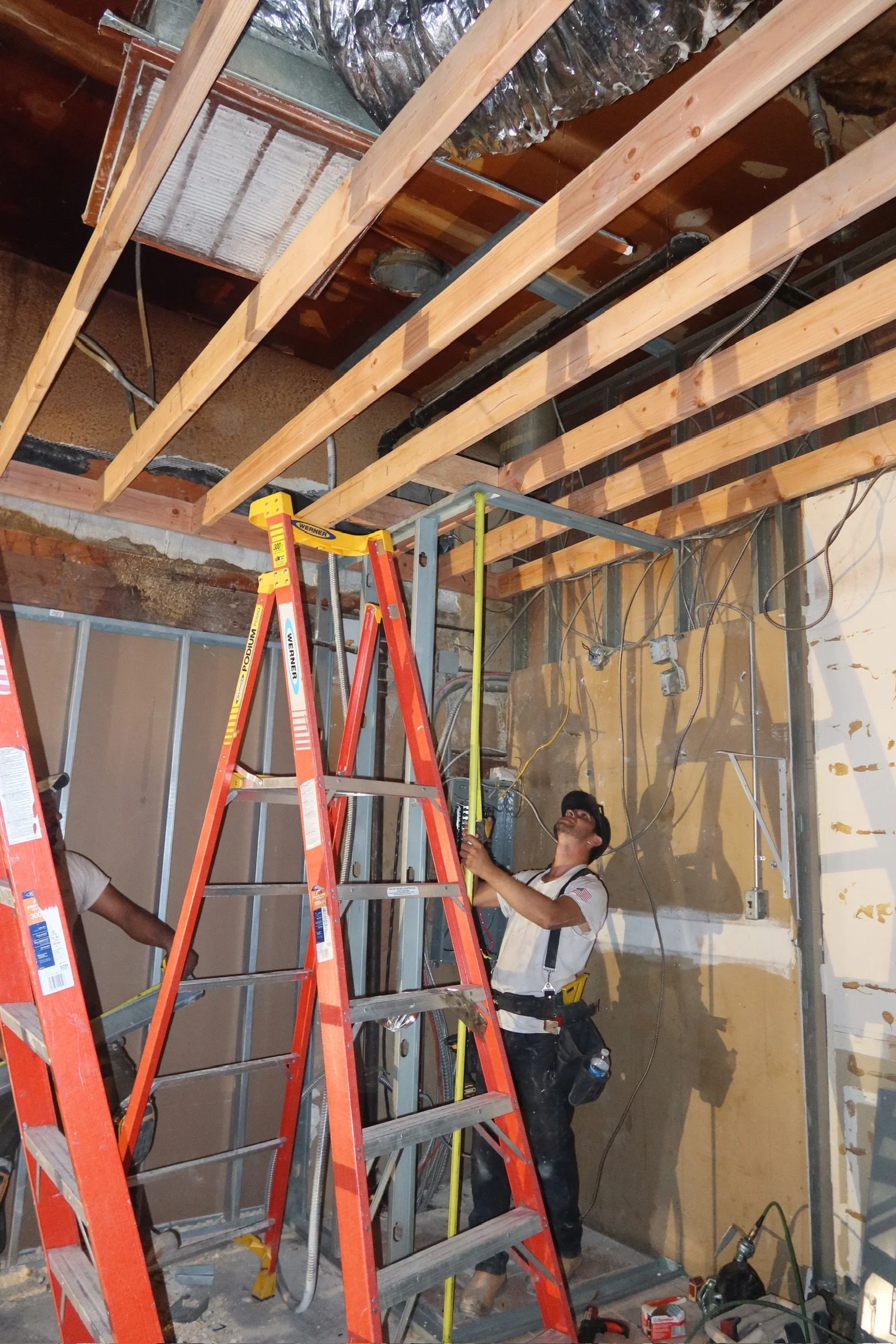 Construction workers installing electrical wiring and framing in a building, with one worker measuring the ceiling height using a tape measure and a ladder in the foreground.