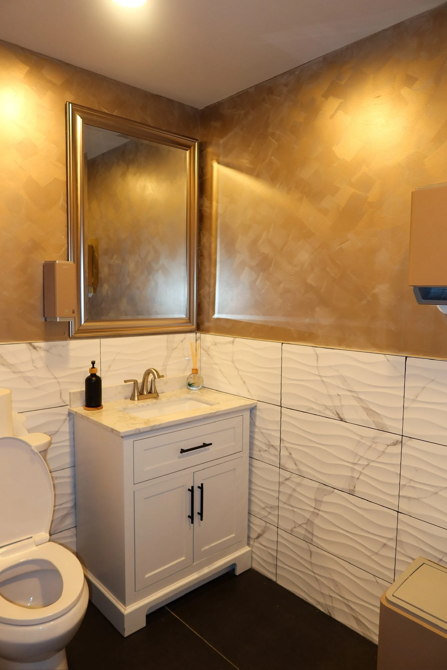 A bathroom with gold metallic upper walls, a large wall mirror, a white vanity with a marble top, a black soap dispenser, a small glass bottle with reed diffusers, and white textured lower wall tiles, with part of a toilet and a beige laundry basket 