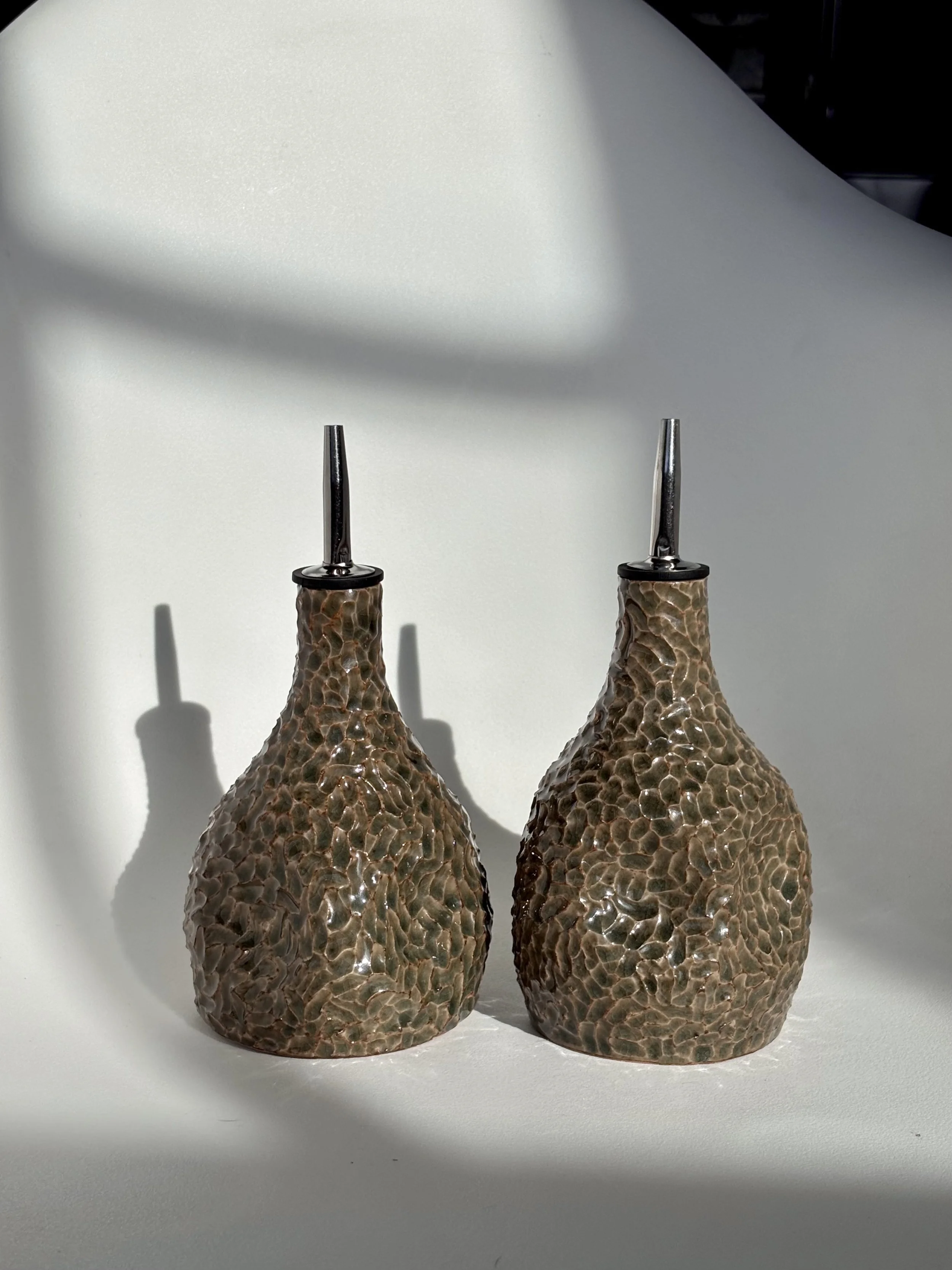 Two decorative glass oil or vinegar dispensers with hammered texture and long metal spouts, placed on a white surface with shadows cast in natural light.