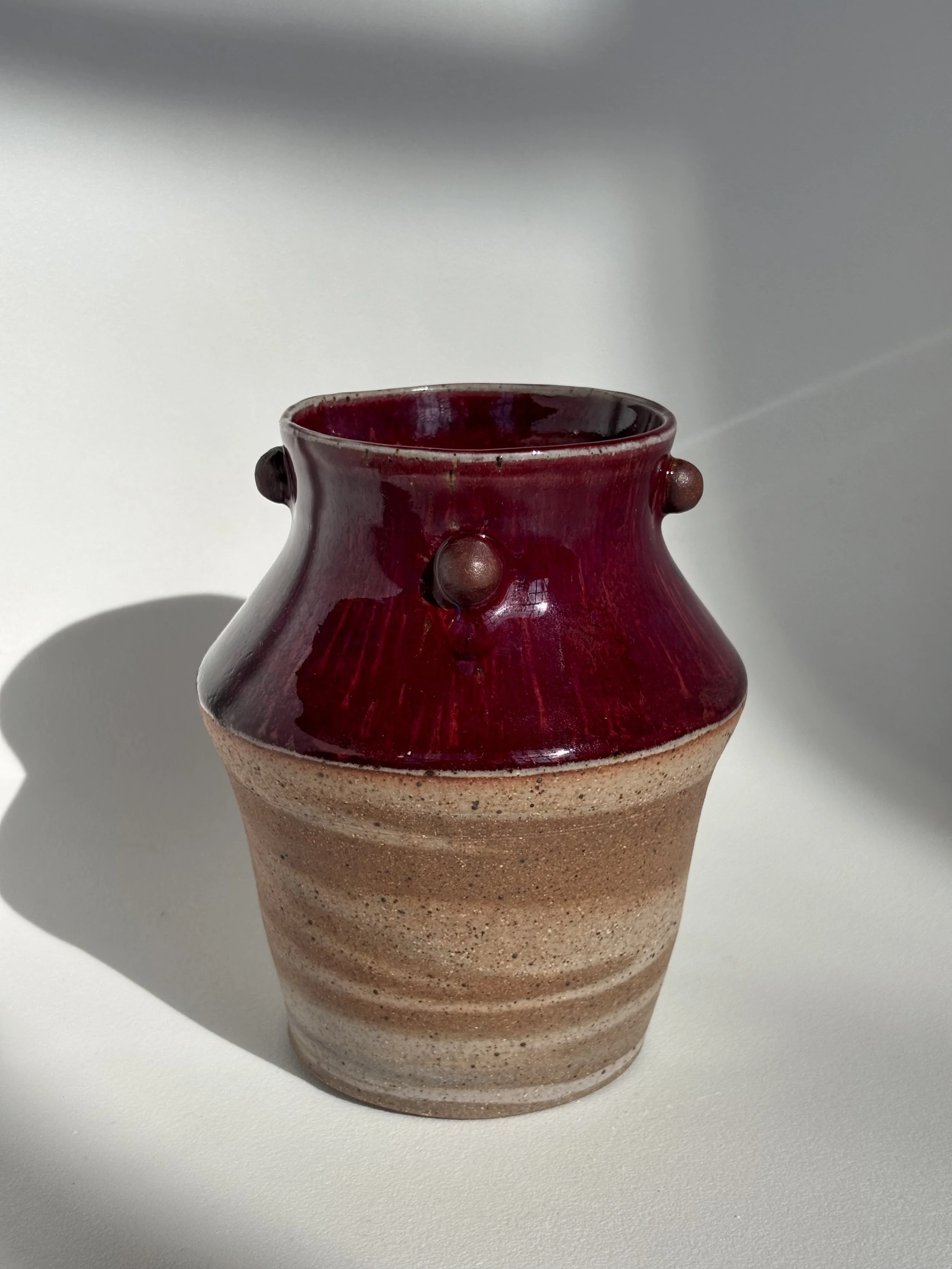 Ceramic vase with a brown and red glaze, featuring small raised knobs around its upper section, placed on a white surface with shadows and lighting highlighting its texture and color.