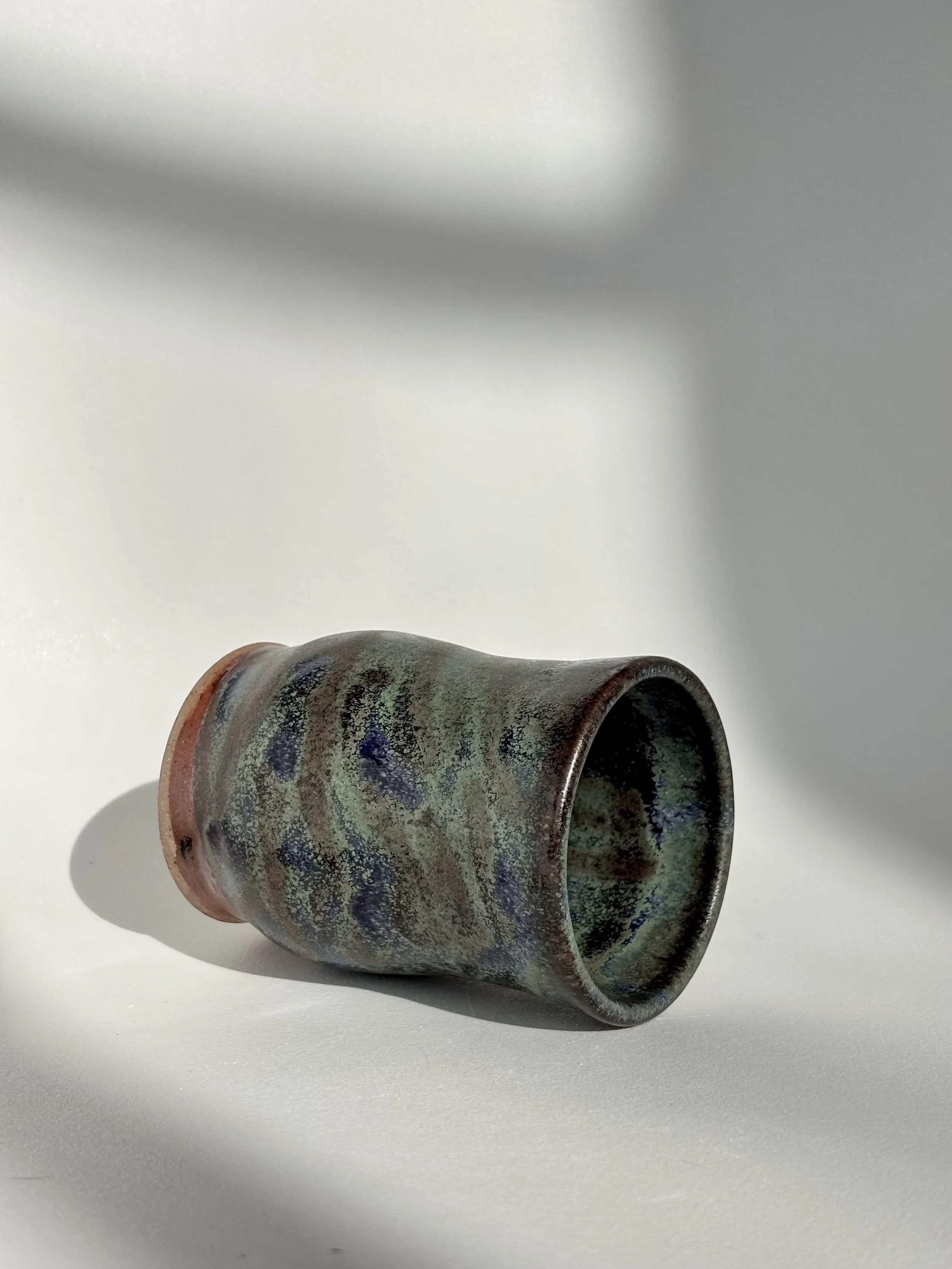 A ceramic cup with a green and brown glaze lying on its side on a white surface, casting a shadow.