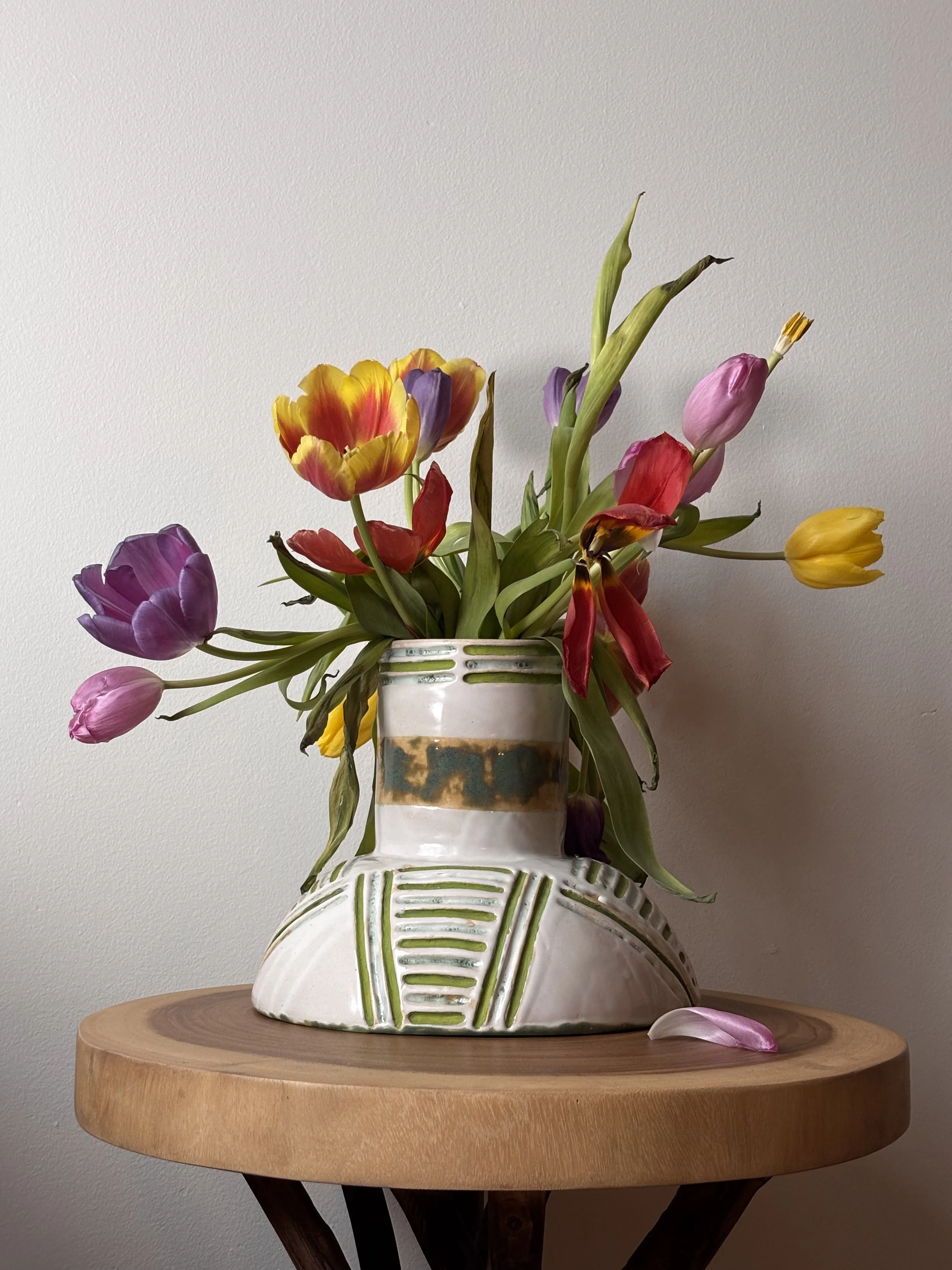 A ceramic vase with green accent lines, filled with colorful tulips, sits on a wooden table against a plain wall.