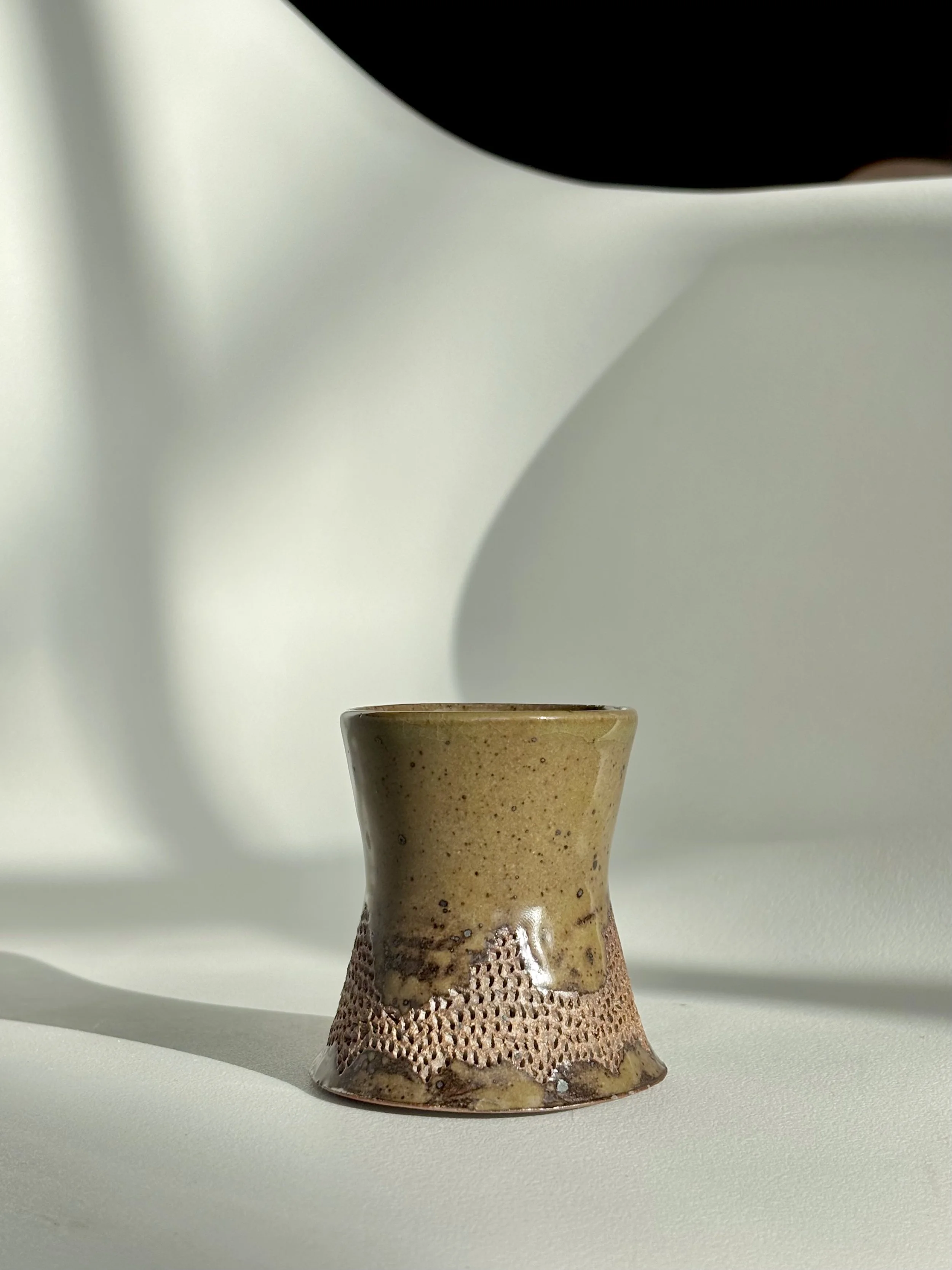 A small ceramic cup with a textured, dotted pattern around the lower part, beige and brown in color, placed on a white surface with abstract shadows in the background.