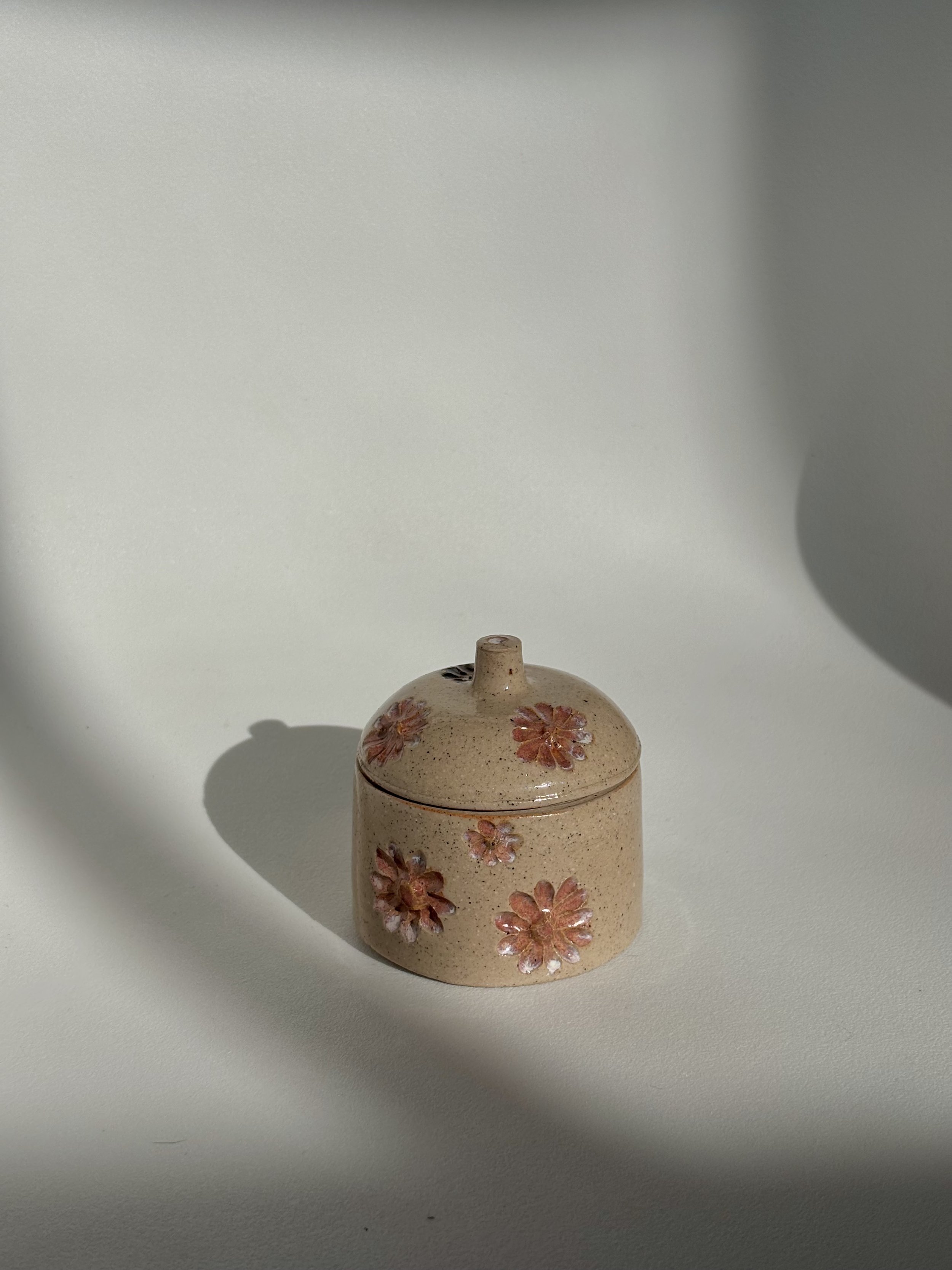 A small ceramic container with a lid, decorated with pink and orange flowers, placed on a white surface with shadows cast around it.