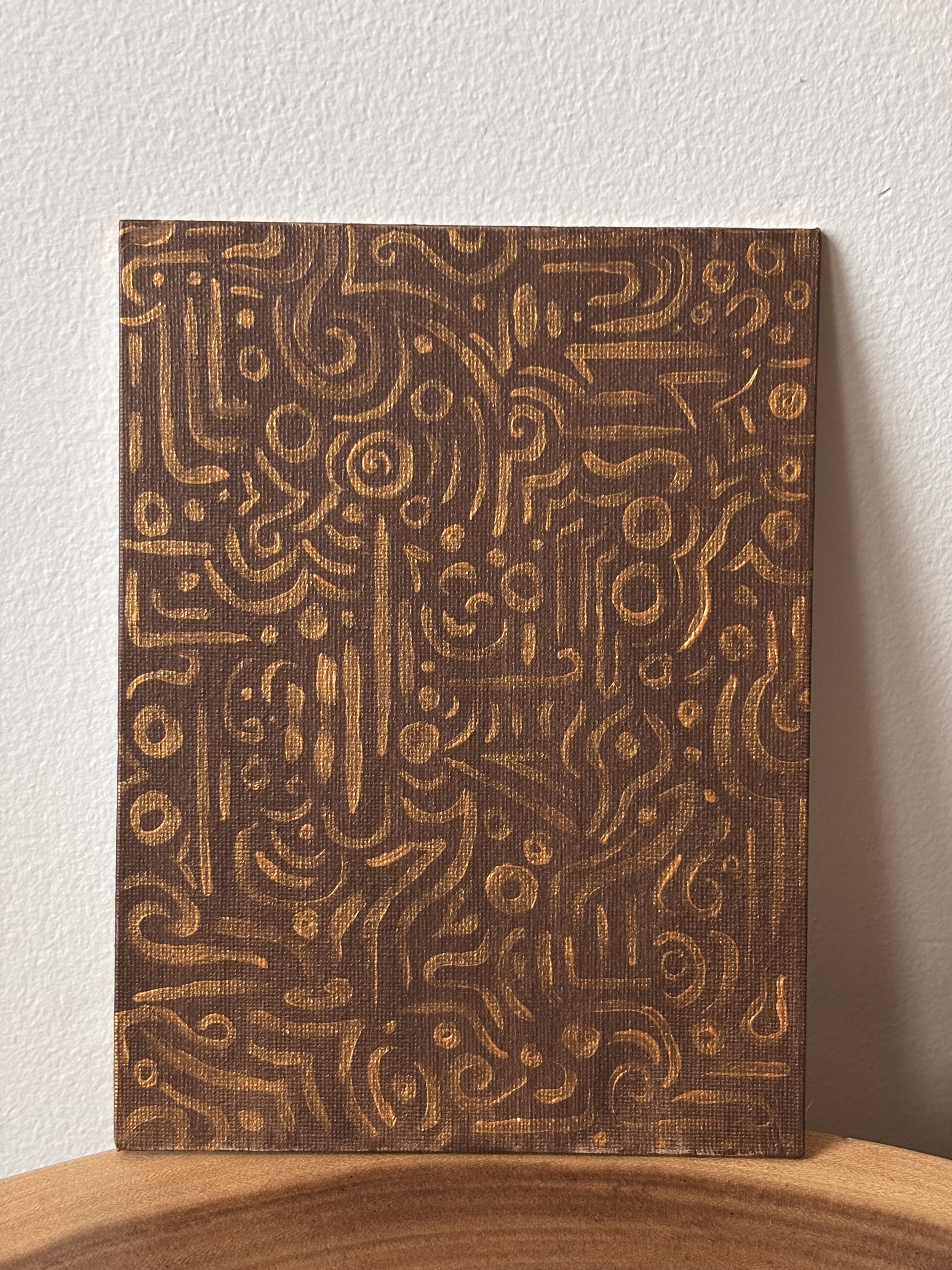 A small canvas with intricate, abstract, gold-colored lines and patterns on a dark brown background, leaning against a plain wall.