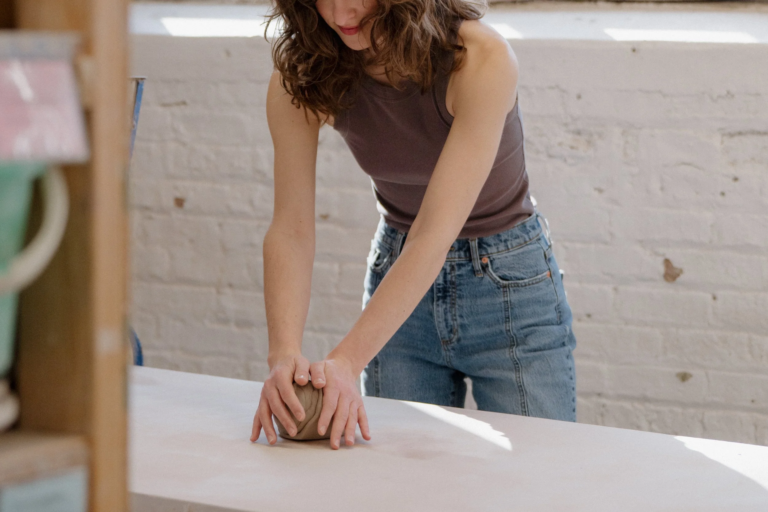 Person kneading clay on a wooden table in a bright room with a white brick wall.