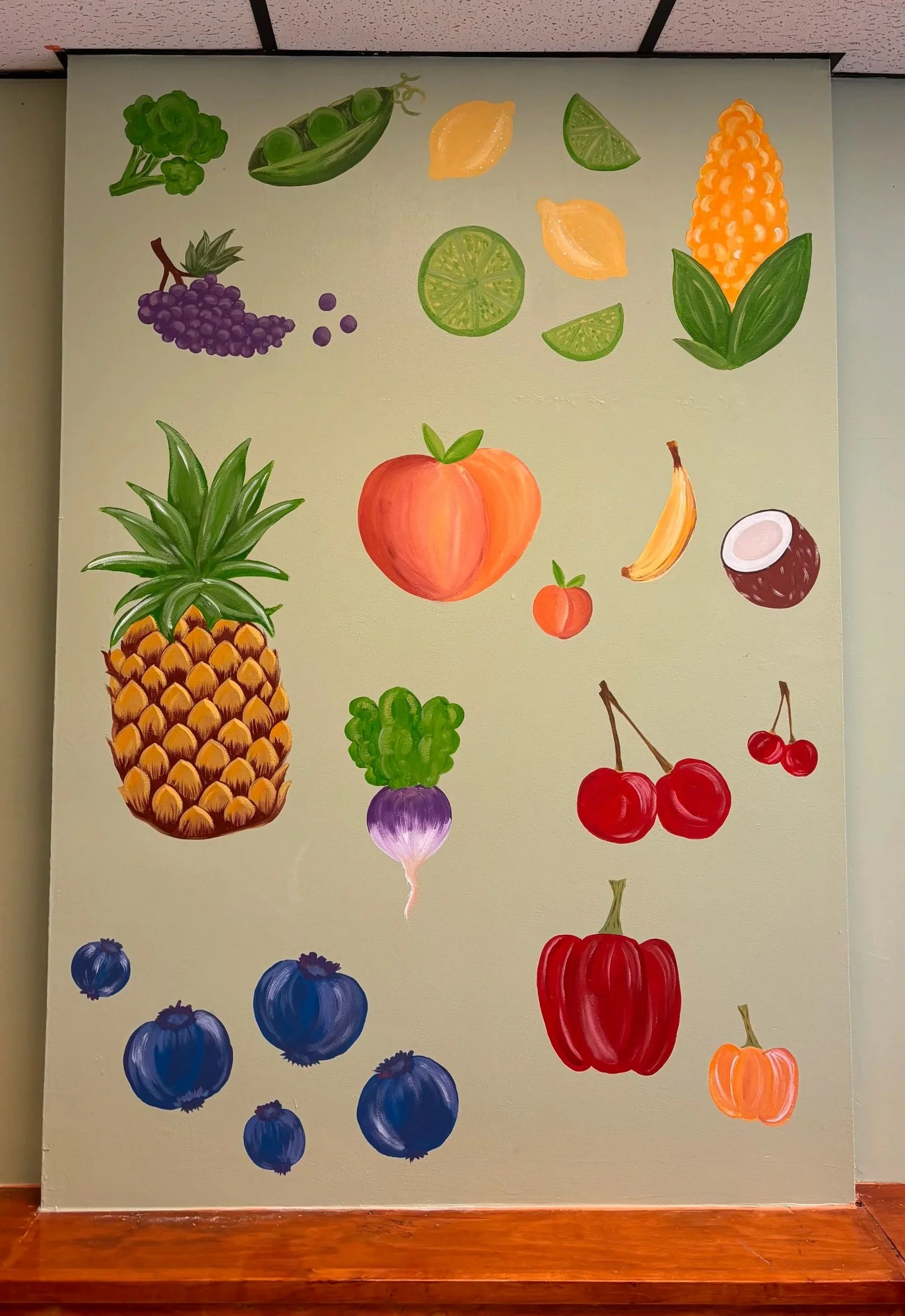 Colorful illustration of various fruits and vegetables on a wall, including broccoli, snap peas, lemons, lime, corn, pineapple, peach, banana, coconut, cherries, blueberries, onion, red bell pepper, and pumpkin.