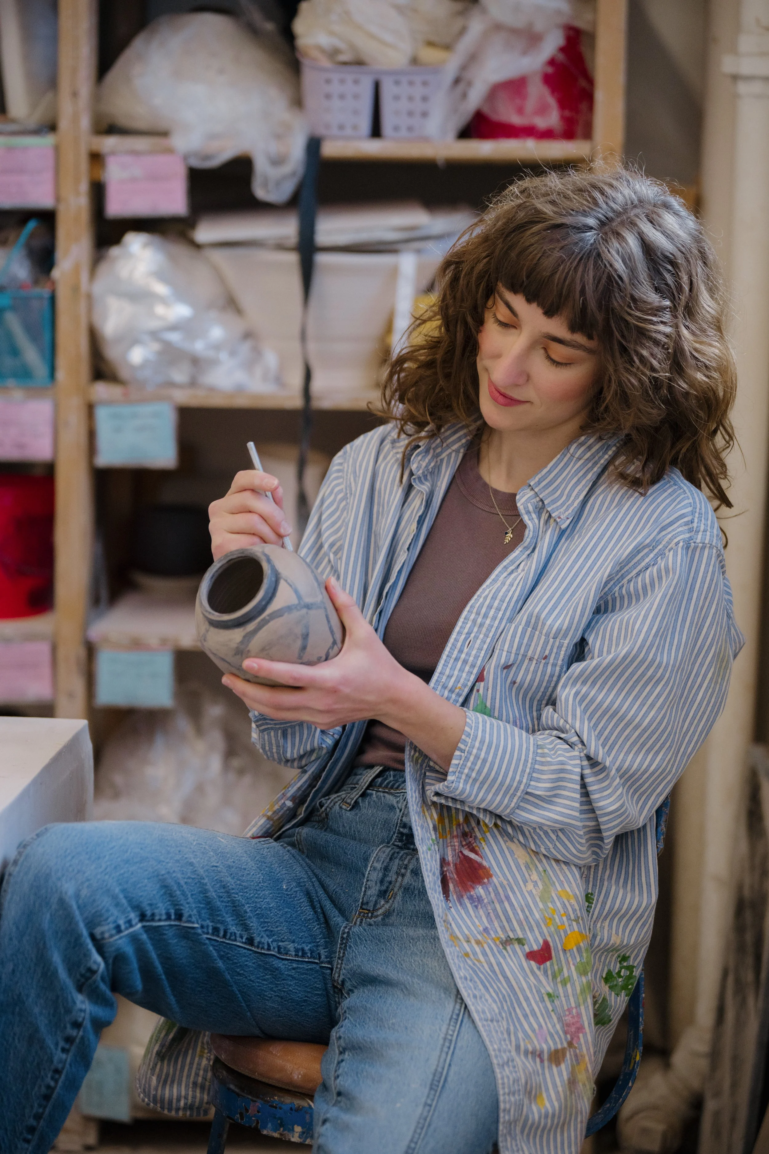 A woman is sitting on a stool in a ceramics studio, painting a ceramic pot with a brush. She has curly brown hair, is wearing a striped shirt over a t-shirt, and jeans. Shelves with supplies and materials are in the background.
