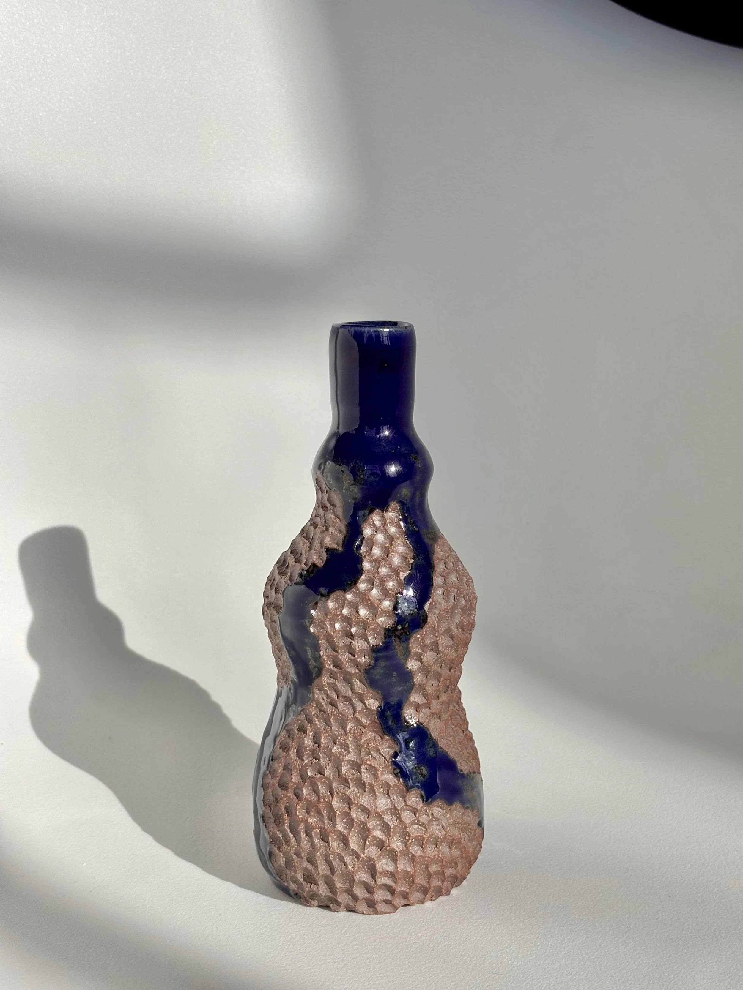 Ceramic vase with textured pinkish-brown surface, dark blue glazed top, and irregular design casting a shadow on a white background.