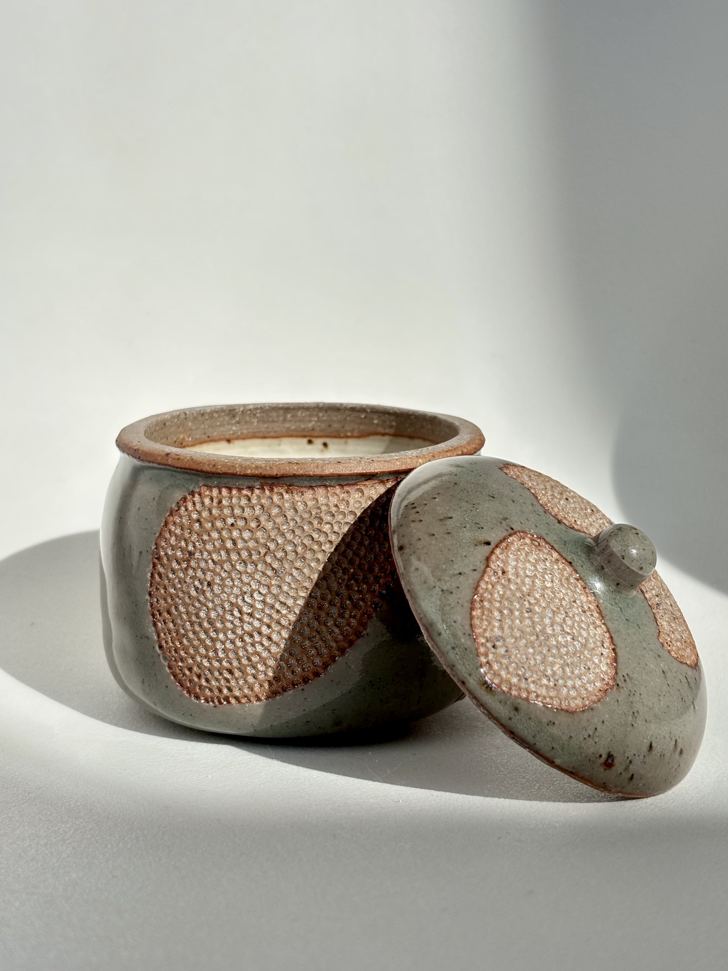 A small ceramic container with a lid, featuring textured pink and gray circular patterns, placed on a white surface with a light background.