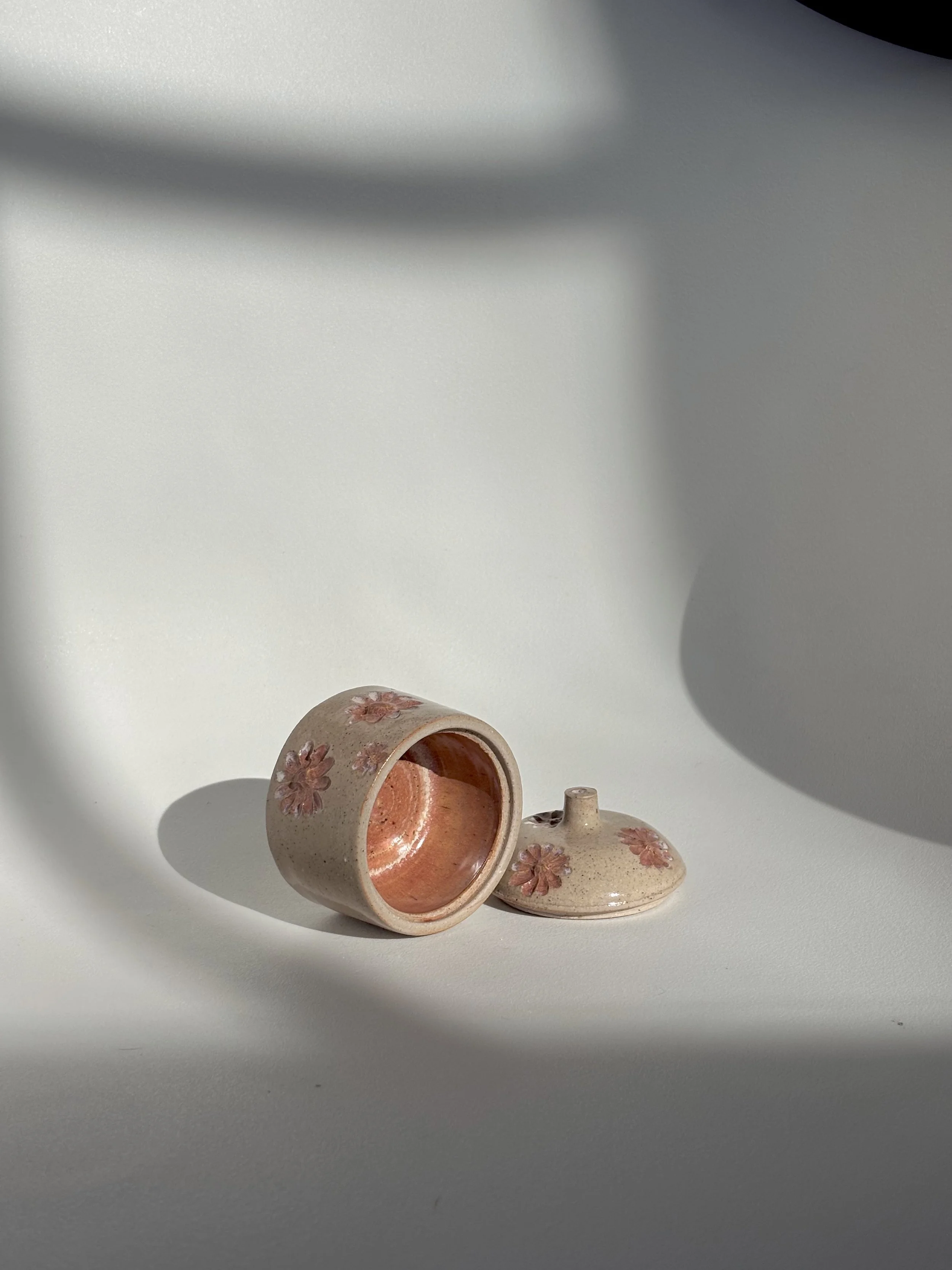 A small ceramic container with pink floral patterns, lying on its side on a white surface with soft shadows.