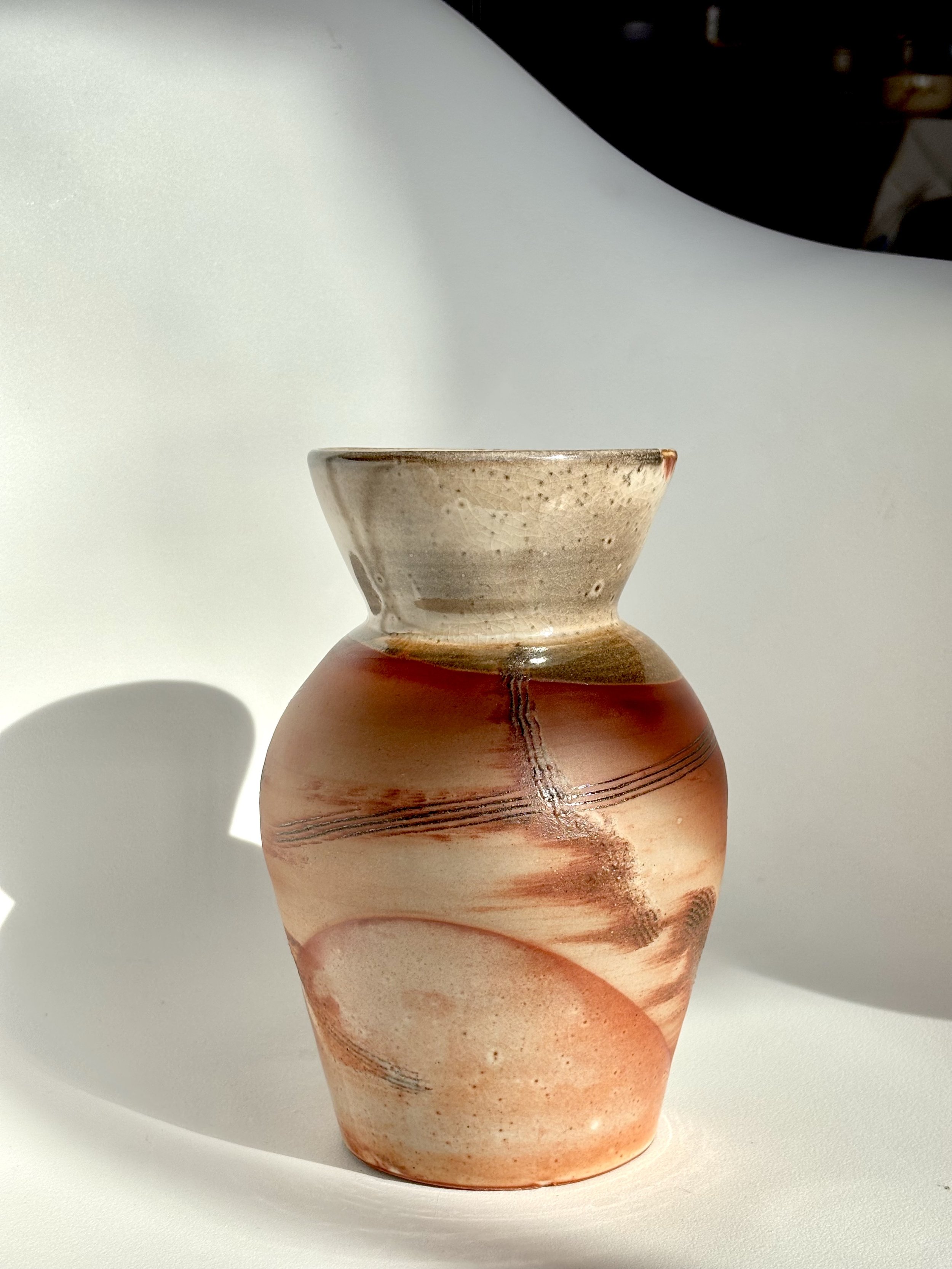 A ceramic vase with earthy tones and abstract swirl patterns against a white background, casting a shadow on the left.