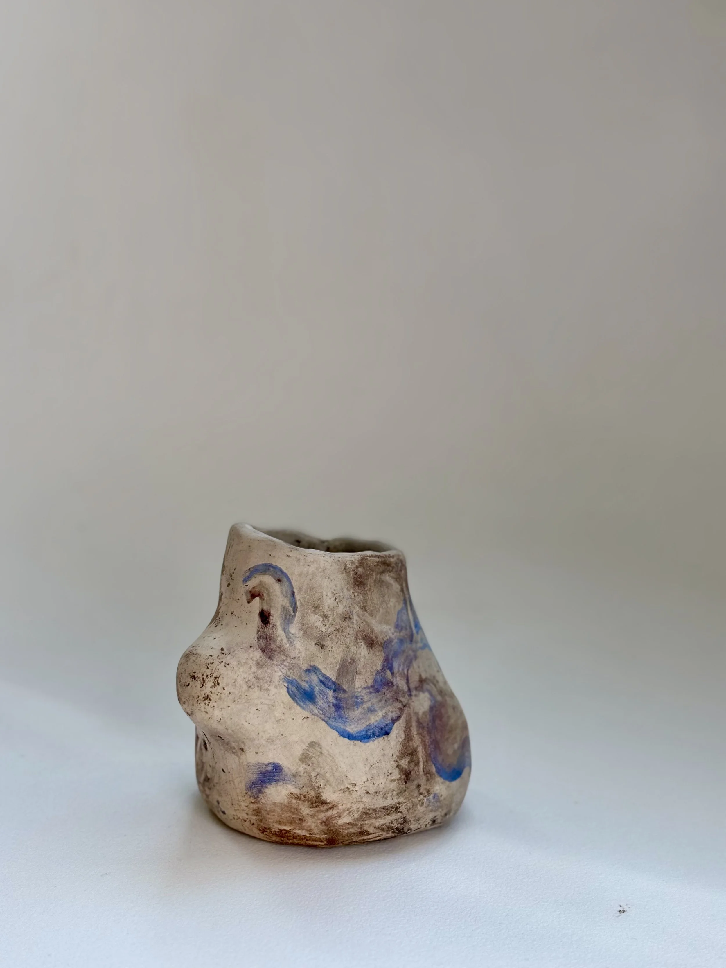 A small, irregularly shaped ceramic vase with blue decorative patterns, placed on a white surface against a plain, light background.