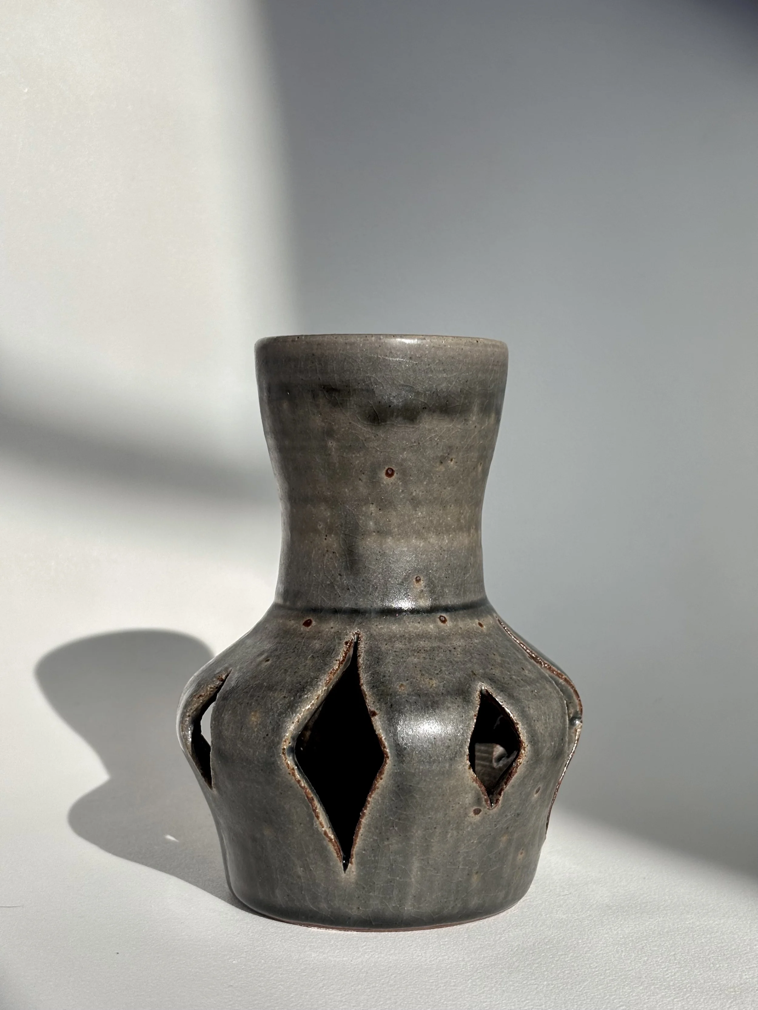 Ceramic vase with a narrow neck, gray glaze, and diamond-shaped cutouts around its body, casting shadows on a white surface.