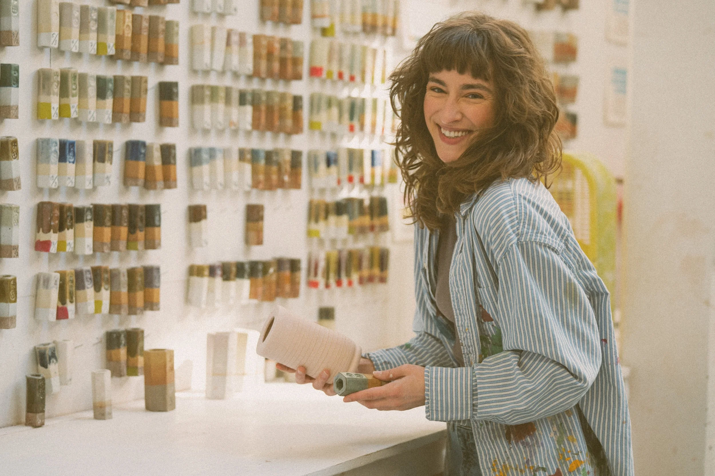 A woman shopping at a paint store, holding a roll of pink painter's tape and a small container of paint, smiling at the camera.