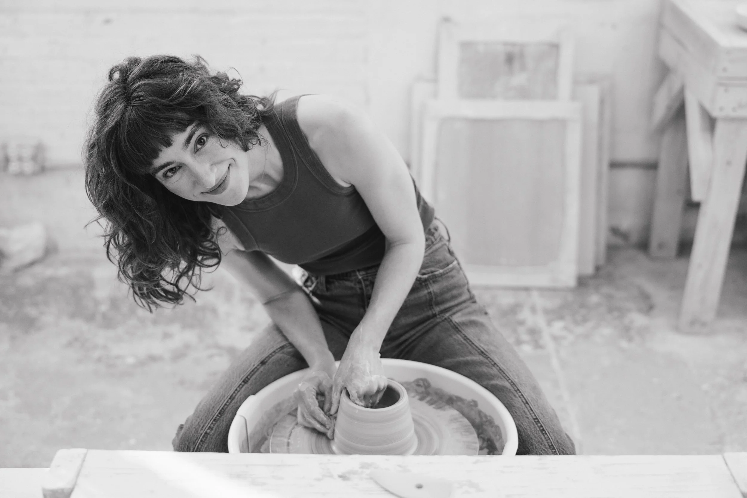 A woman with curly hair and a dark tank top is leaning over a pottery wheel, shaping clay with her hands, while smiling at the camera in a pottery studio.