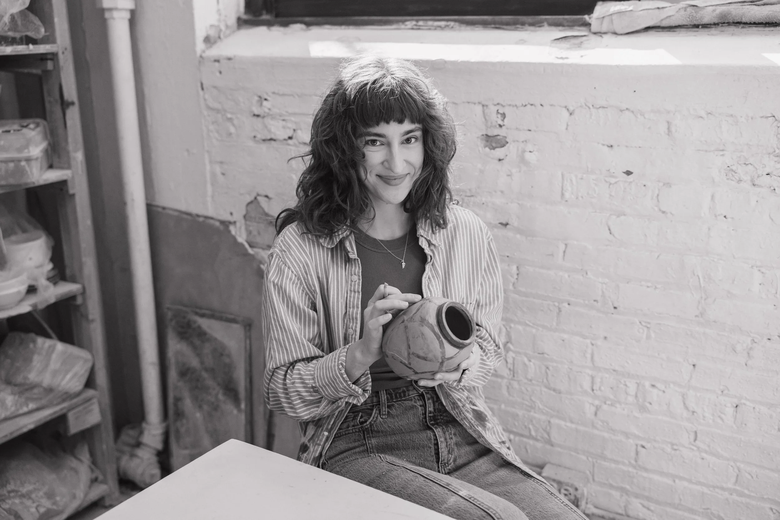 A young woman sitting in a studio or workshop holding a ceramic vase and smiling at the camera. She has curly hair, wearing a striped shirt and jeans, with shelves containing pottery and art supplies in the background.