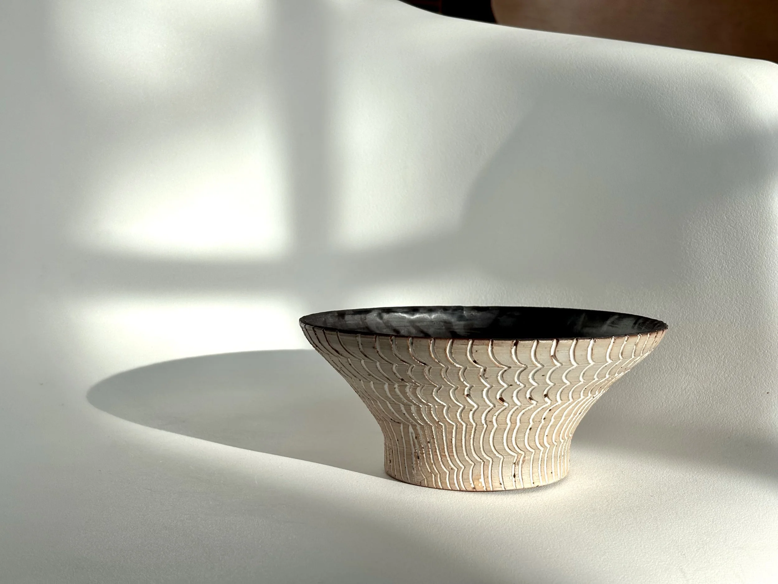 A decorative ceramic bowl with a textured exterior and dark interior, placed on a white surface with shadows cast by sunlight.