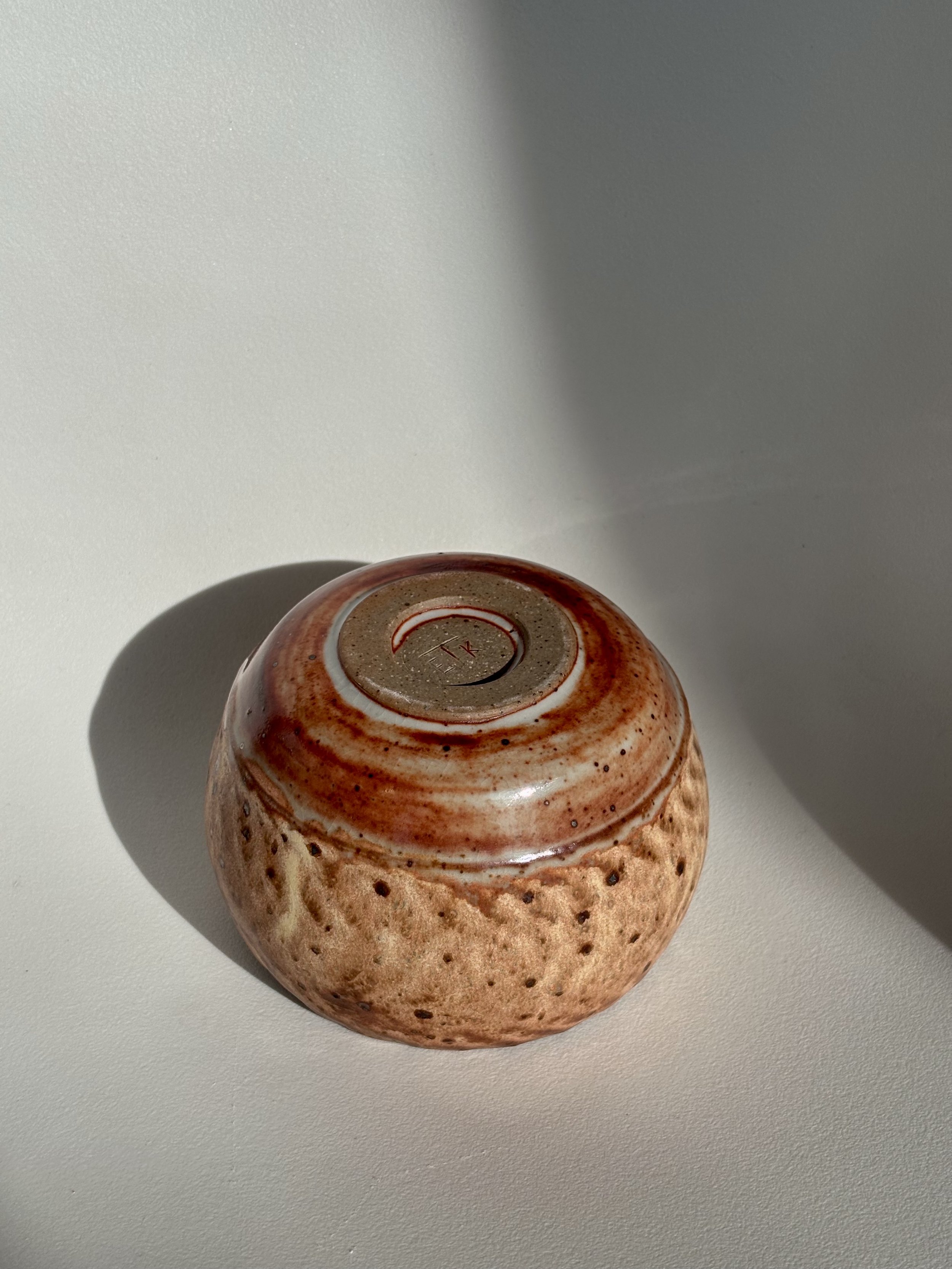 A small, round, speckled ceramic cup with a reddish-brown glaze, viewed from the bottom with an unglazed circular foot and a faint mark inside the foot, placed on a white surface with a shadow cast to the left.