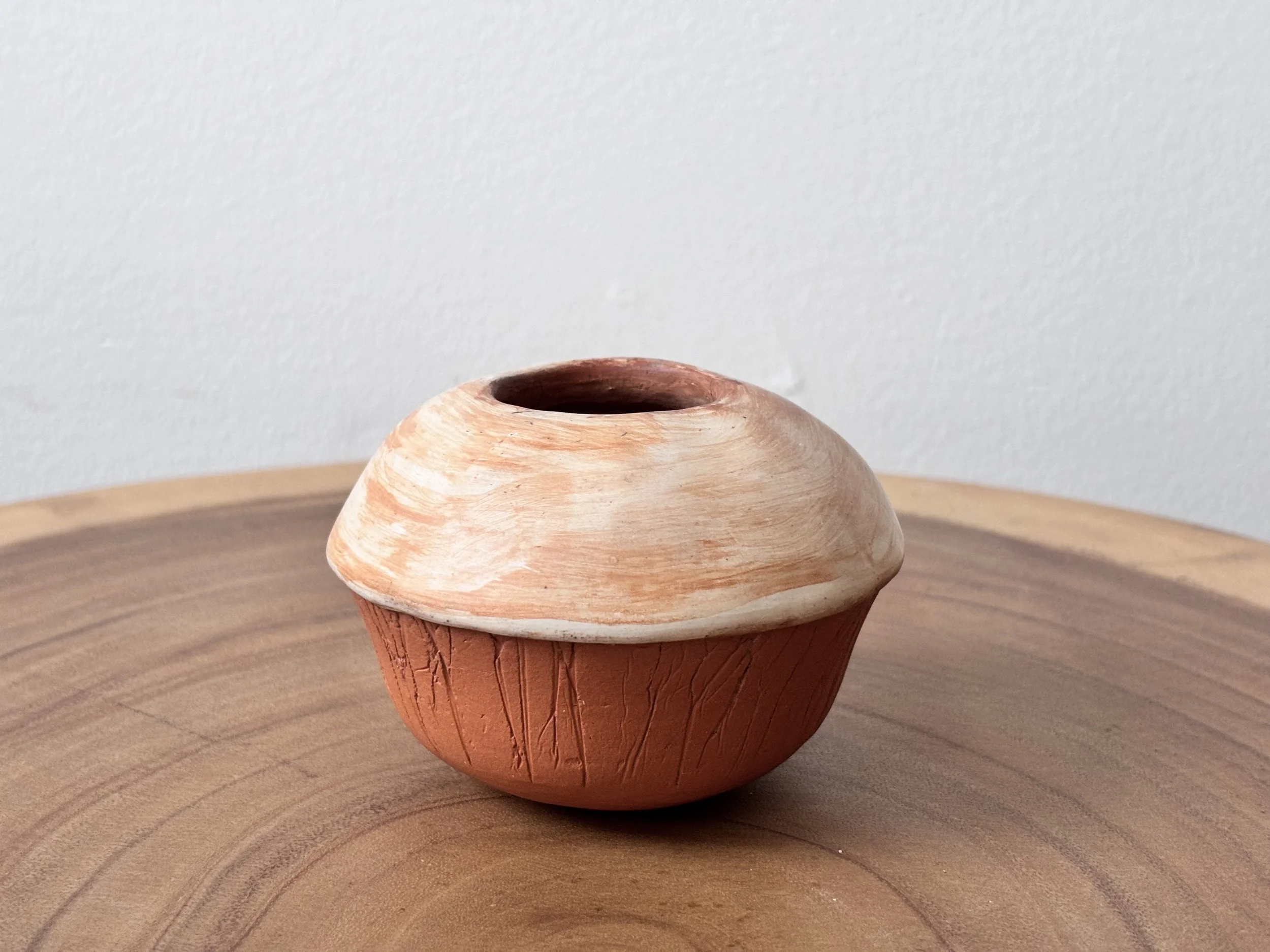 A small round pottery vessel with a wide opening on top, a textured reddish-brown bottom, and a beige, slightly domed lid, sitting on a wooden surface against a plain white background.