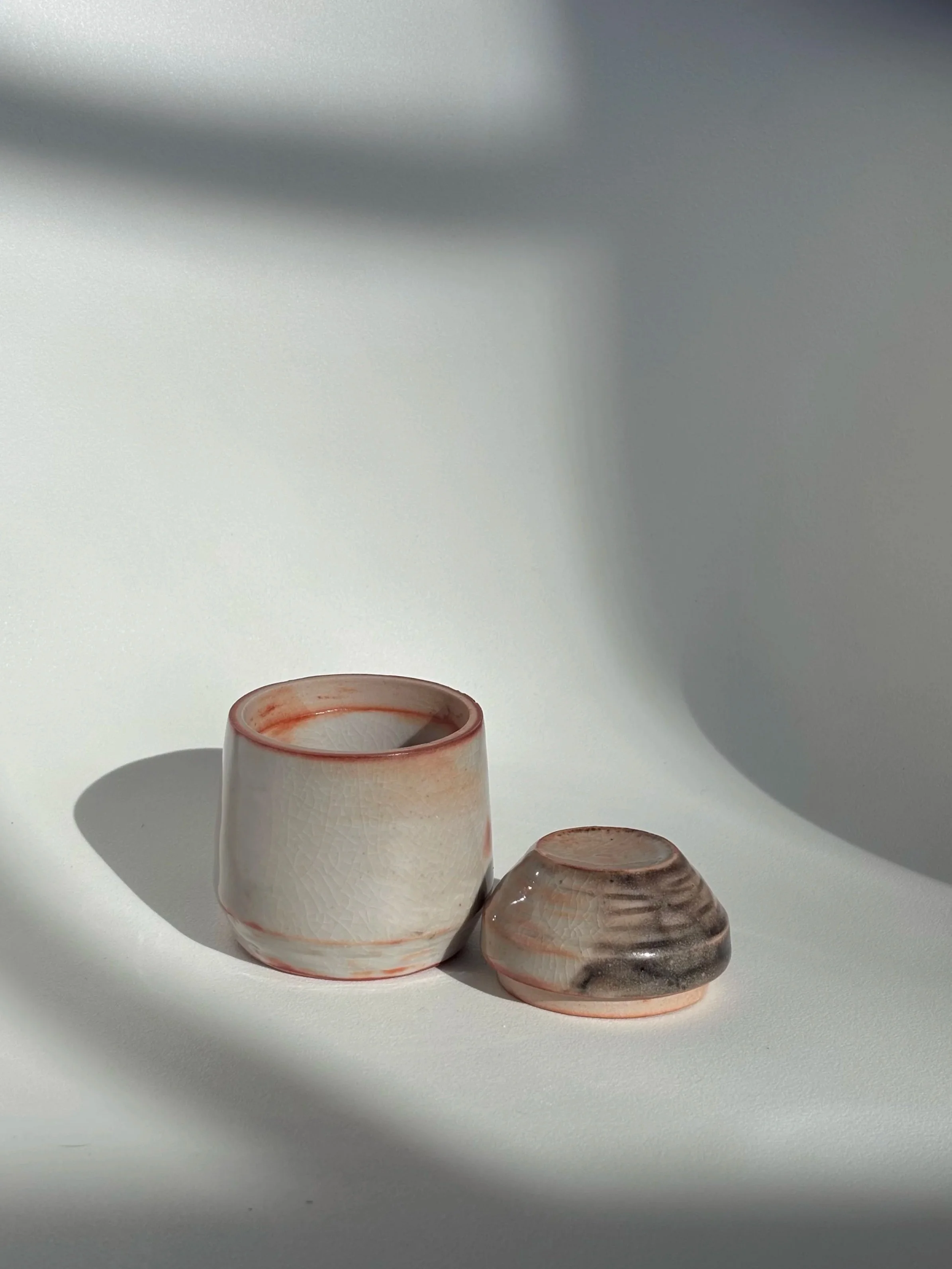 Two small ceramic containers with a crackle glaze, one is rounded and the other is slightly flattened, placed on a white surface with shadows and soft lighting.