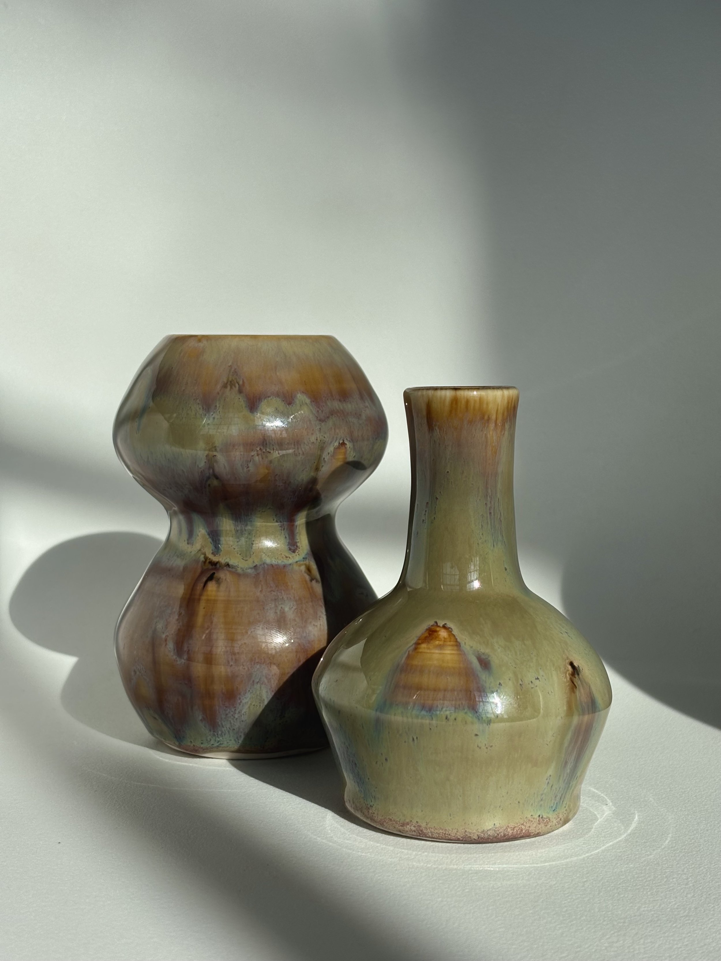 Two decorative vases with abstract, colorful glaze finishes on a white surface with shadows.