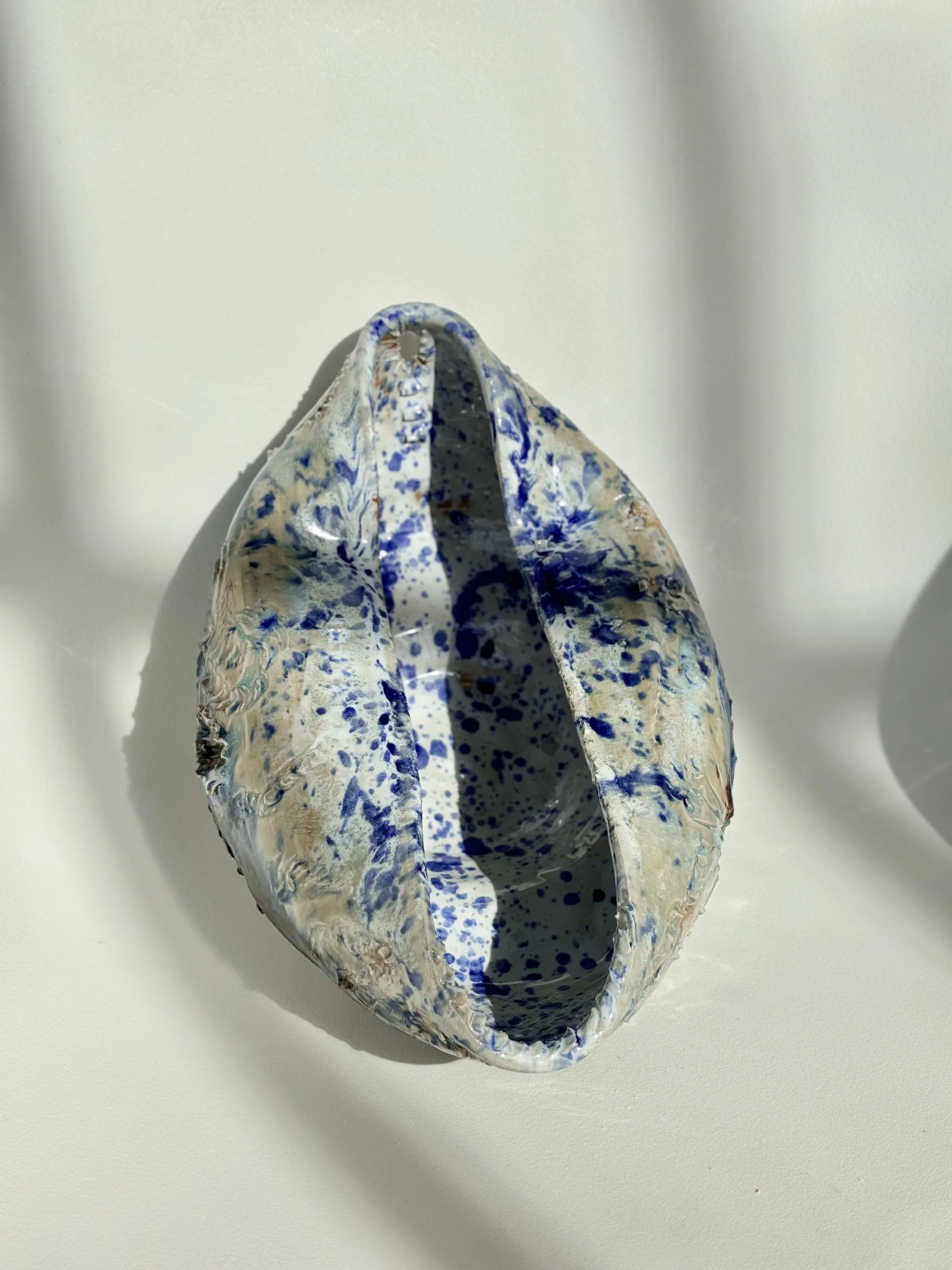 Ceramic vessel with a blue and beige splattered glaze, open at the top, casting a shadow on a white background.