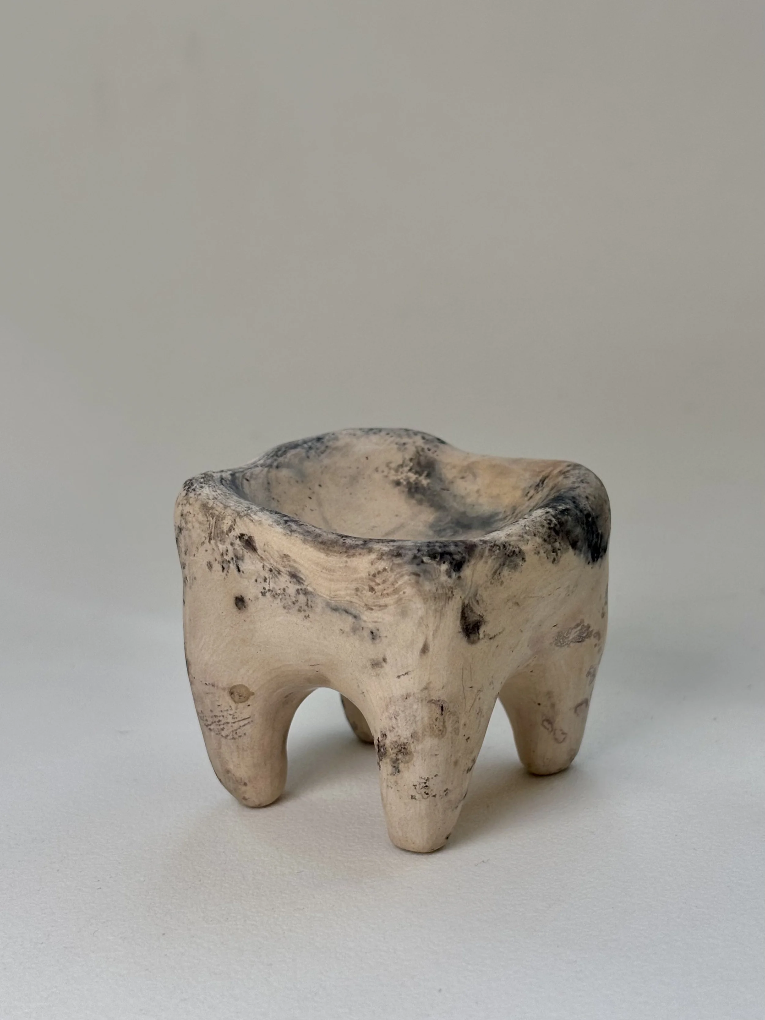 Small carved tooth-shaped object made from light-colored clay with dark spots, placed on a plain light surface.