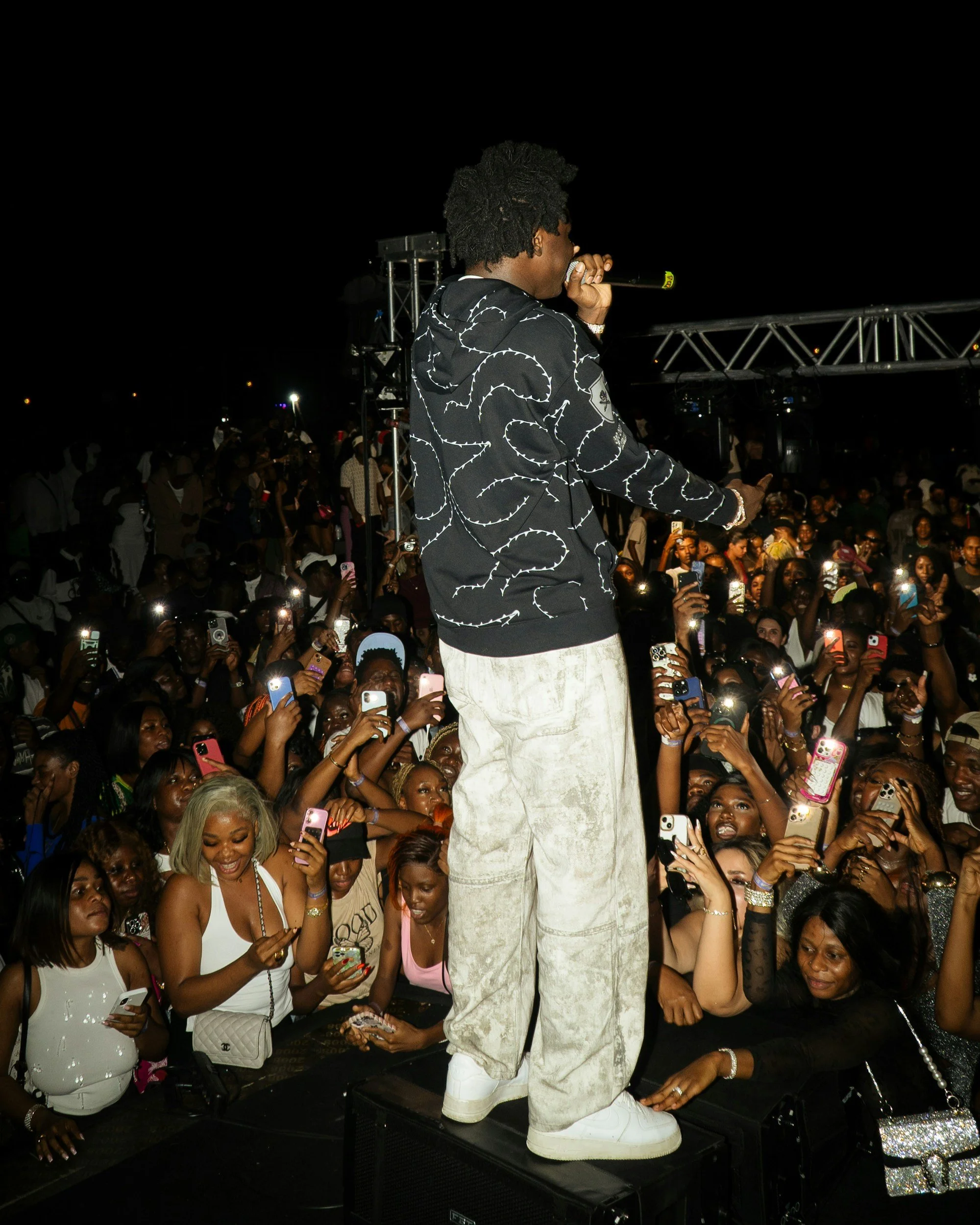 A male performer on stage at night, holding a microphone, facing a large crowd of fans who are taking photos and videos with smartphones.