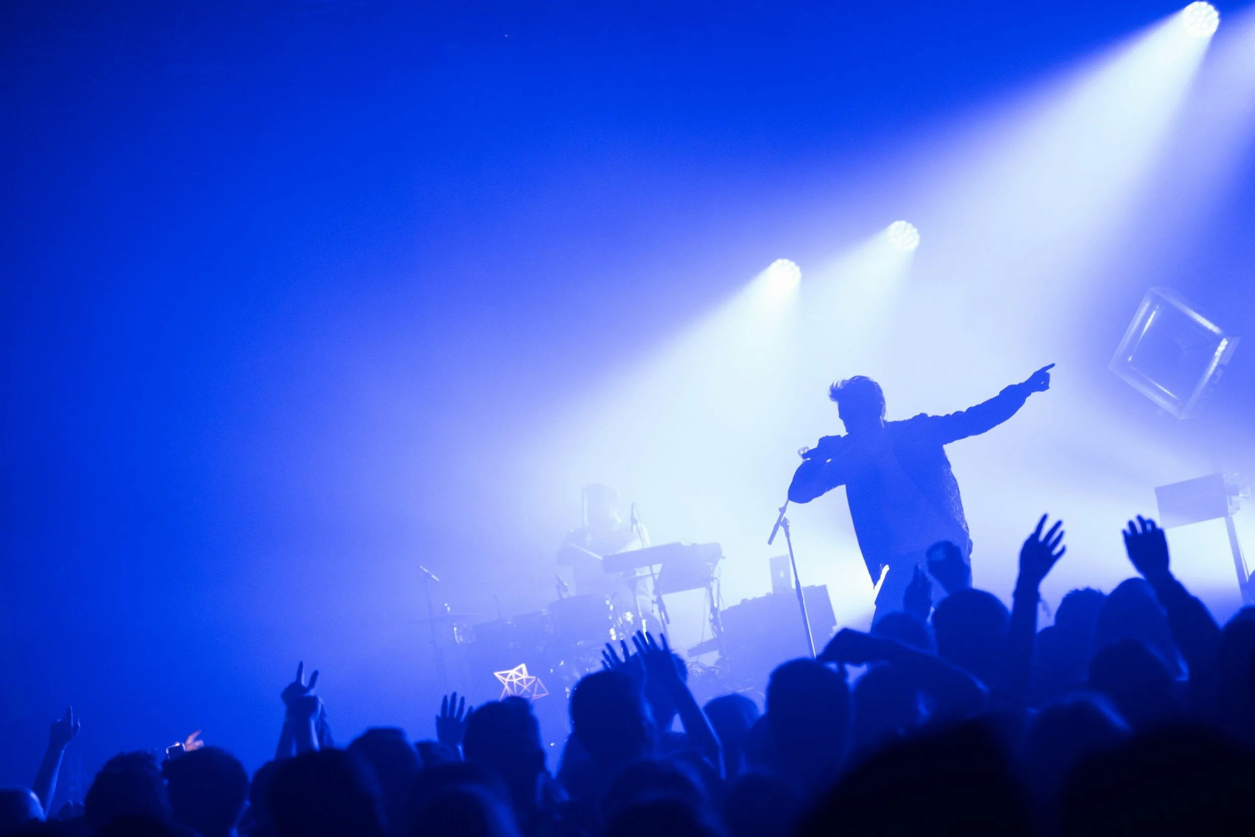 A silhouette of a singer with a microphone performing on stage with bright blue lights and audience raising their hands.