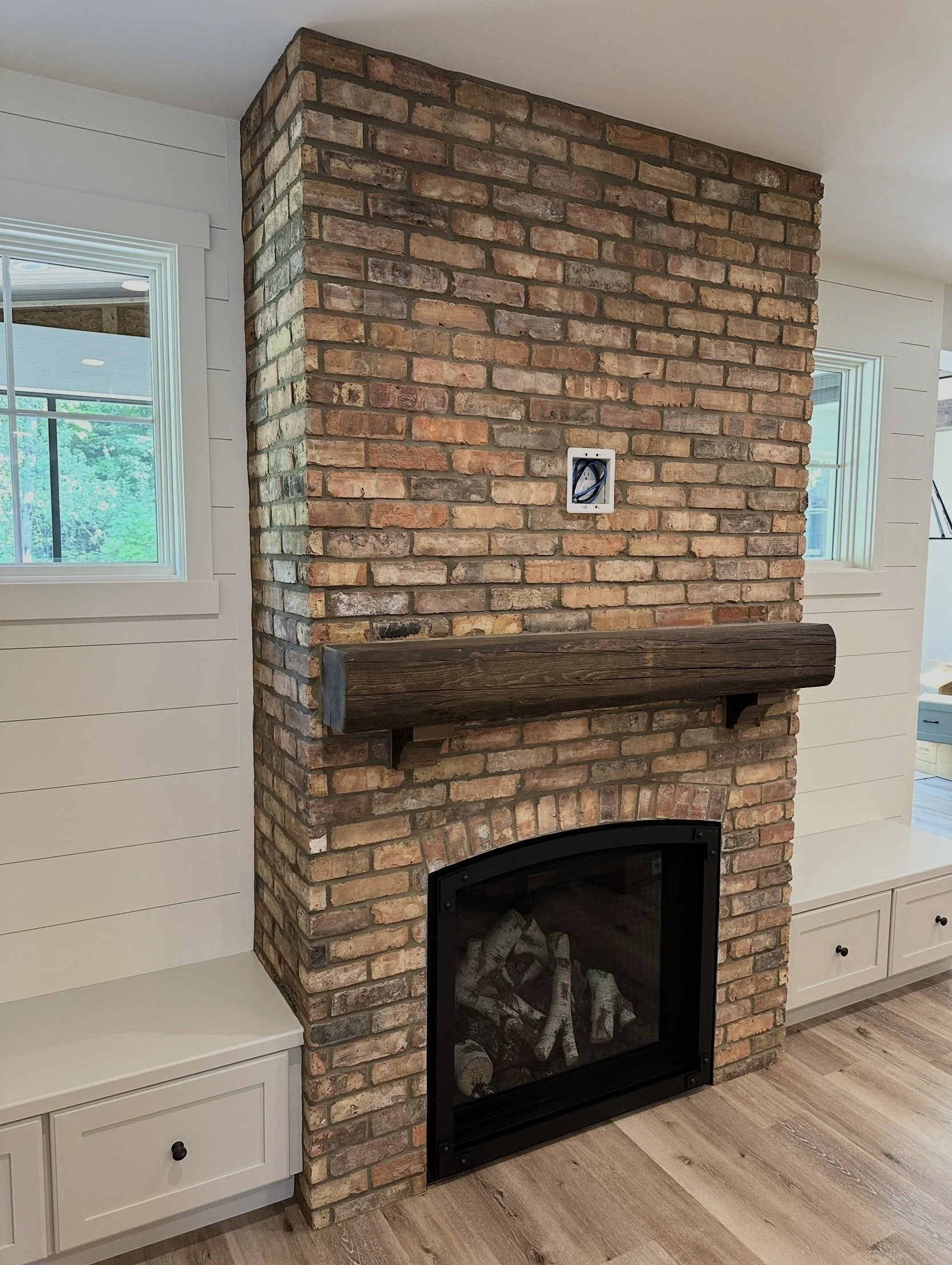 Interior view of a brick fireplace with a wooden mantel, adjacent white built-in cabinets, and windows on each side showing greenery outside.