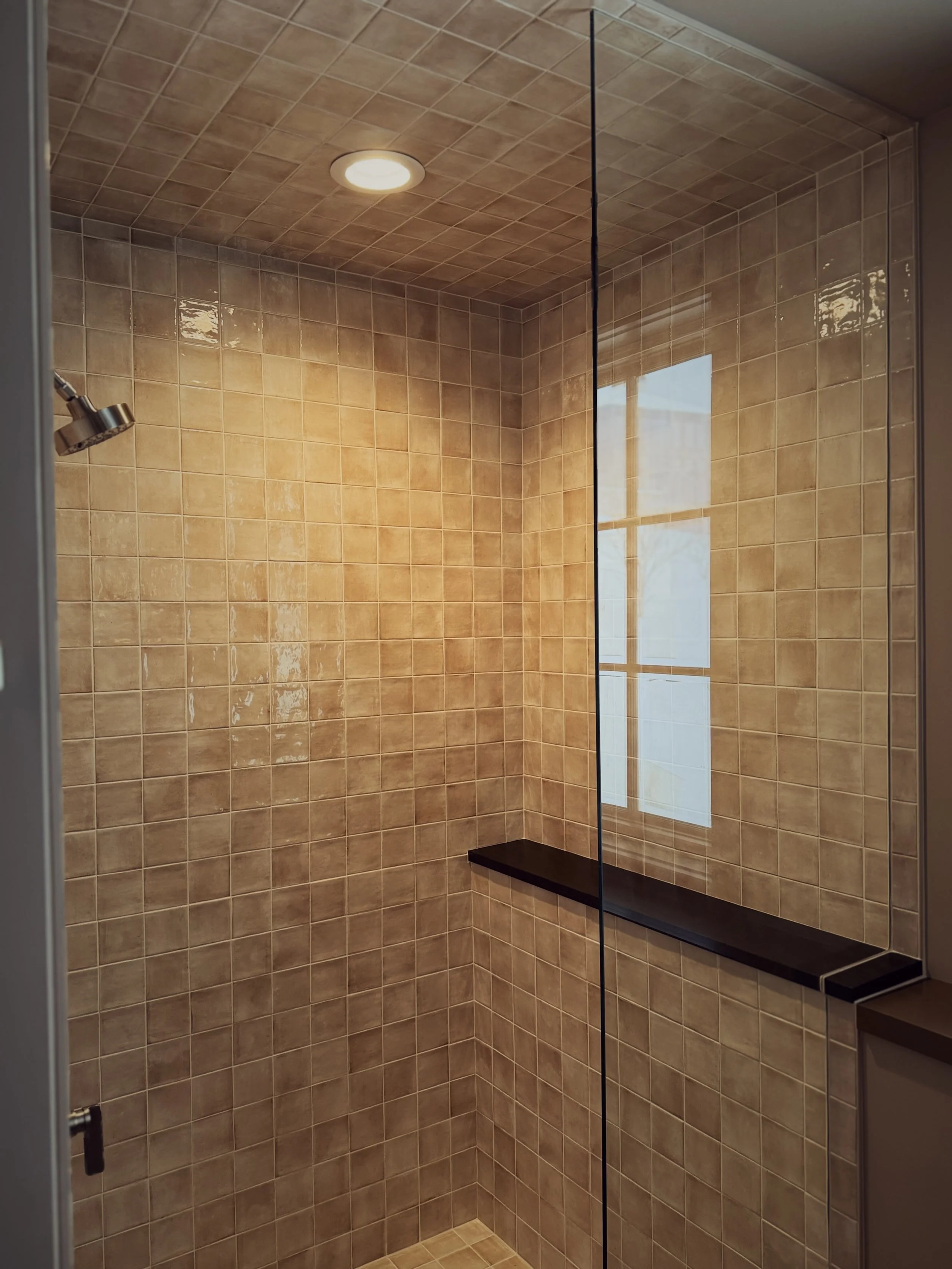 View of a shower with beige tiled walls, a black shelf, a ceiling light, a glass door, and a window reflecting outside scenery.