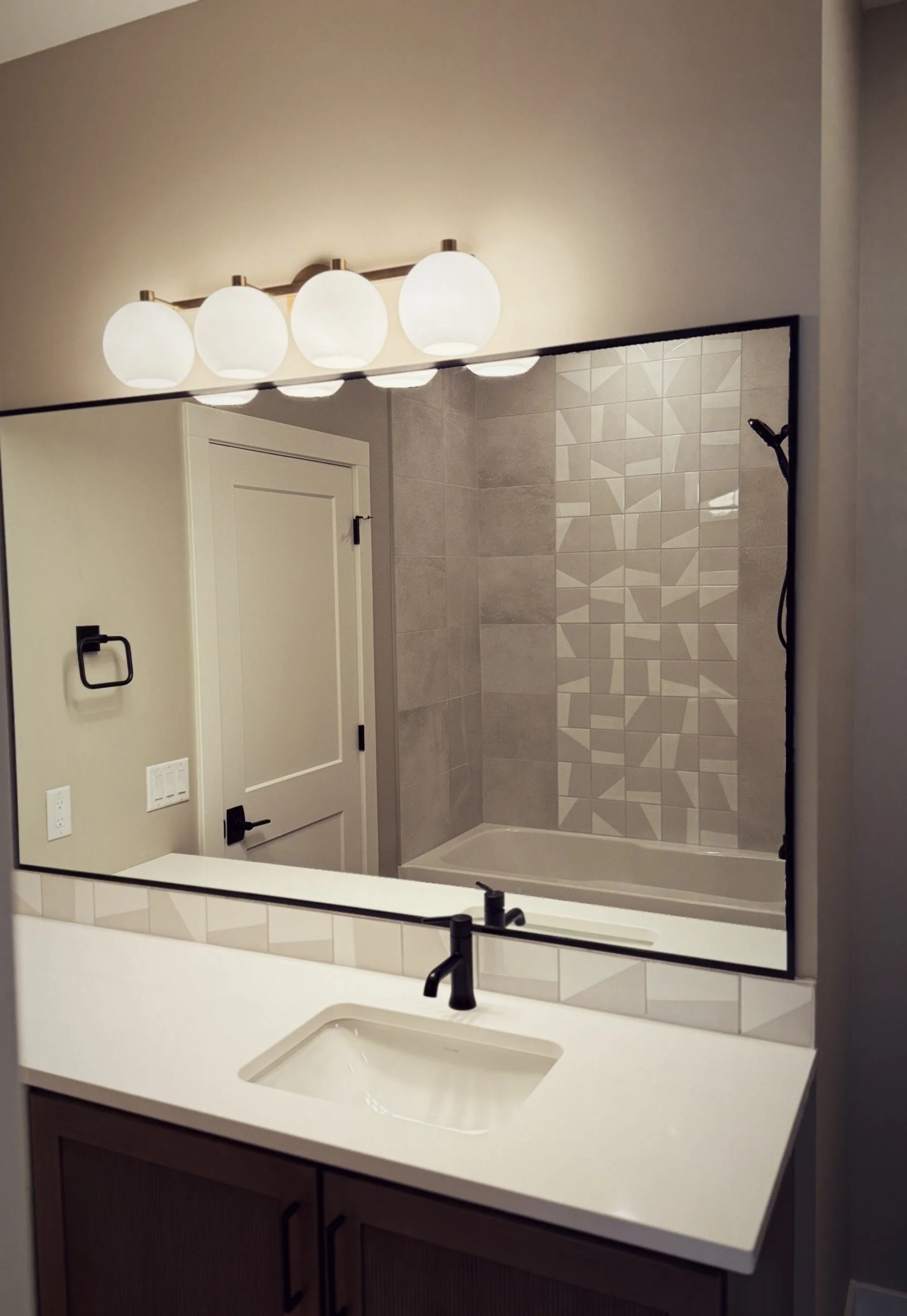 A modern bathroom mirror reflecting a shower area with geometric patterned tiles, a white bathtub, a white door, a beige wall, and a row of four globe-shaped light fixtures above the mirror.