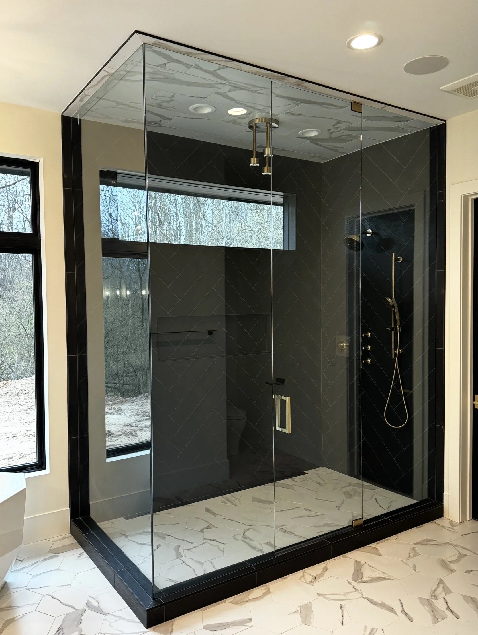 Modern glass-enclosed shower with black tiles and marble tiles on the floor next to a window showing an outdoor wooded landscape.