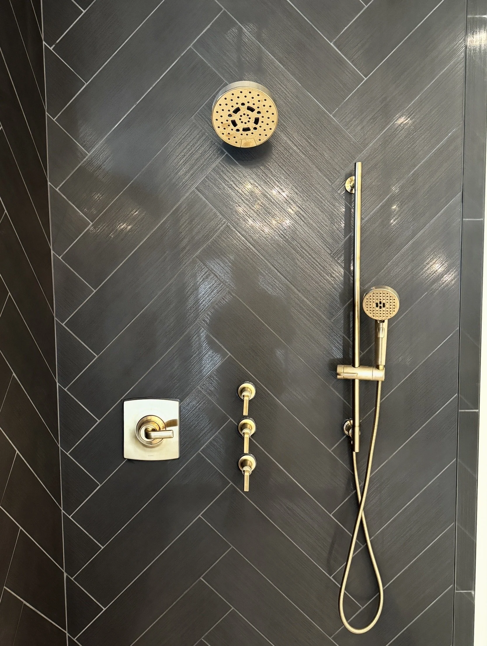 Black tile shower wall with gold fixtures, including a showerhead, handheld shower, and controls.
